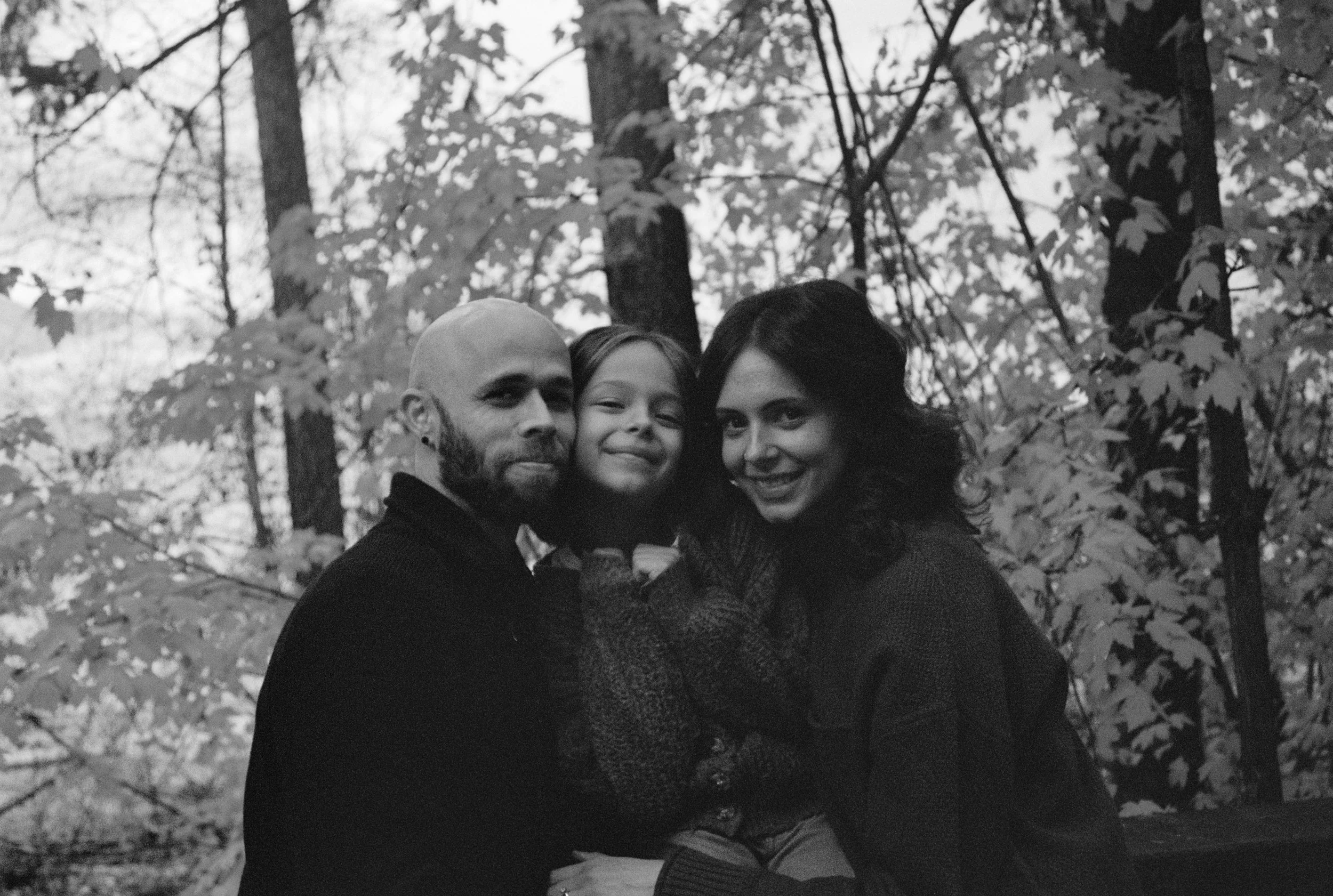 A family photo of a man, woman, and young girl sitting closely together outdoors in a wooded area.