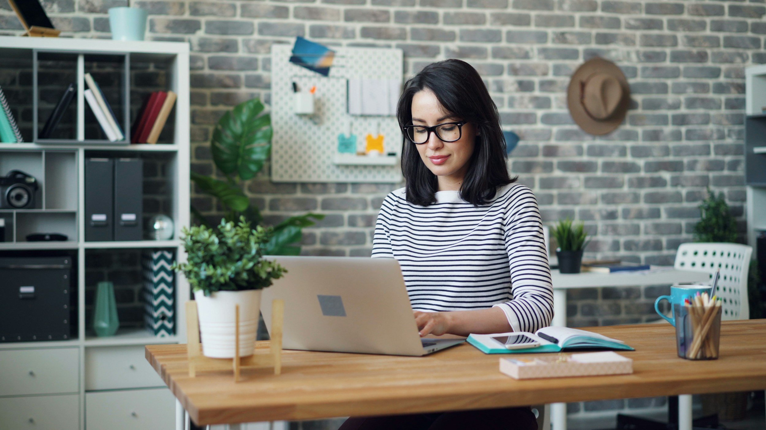 A woman with black hair, glasses, and a striped shirt working on a laptop at a wooden desk in a modern office with plants and shelves.