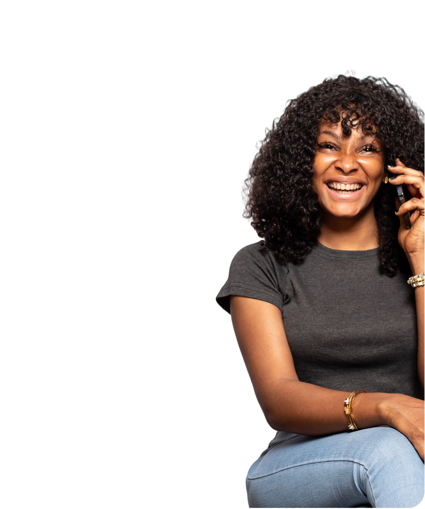 A smiling woman with curly black hair, wearing a dark gray t-shirt and jeans, talking on a cell phone.
