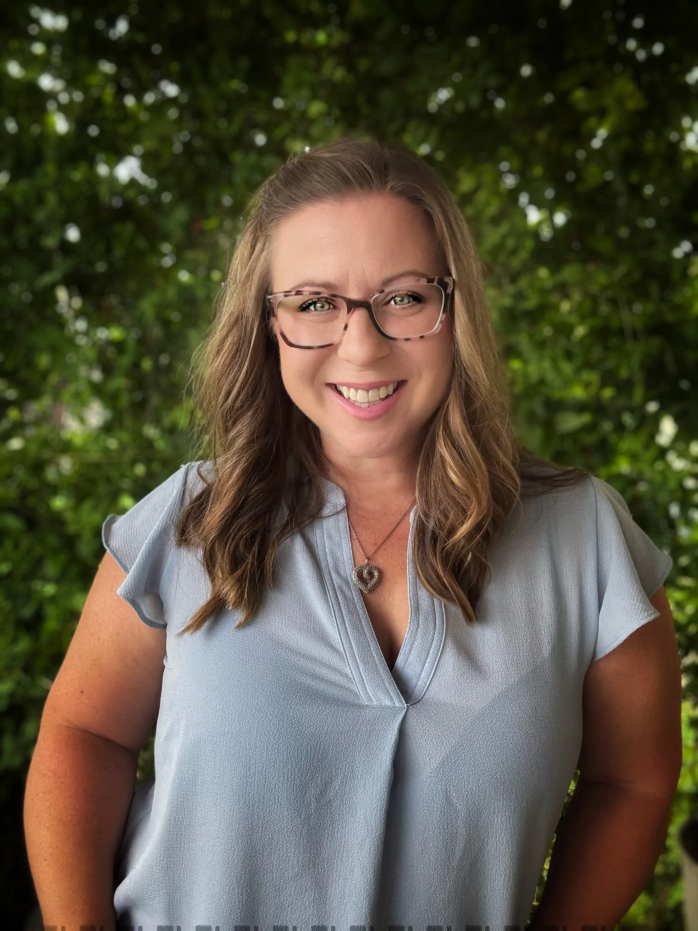 Smiling woman with glasses, wearing a light blue blouse and a heart-shaped necklace, standing outdoors against a green leafy background.