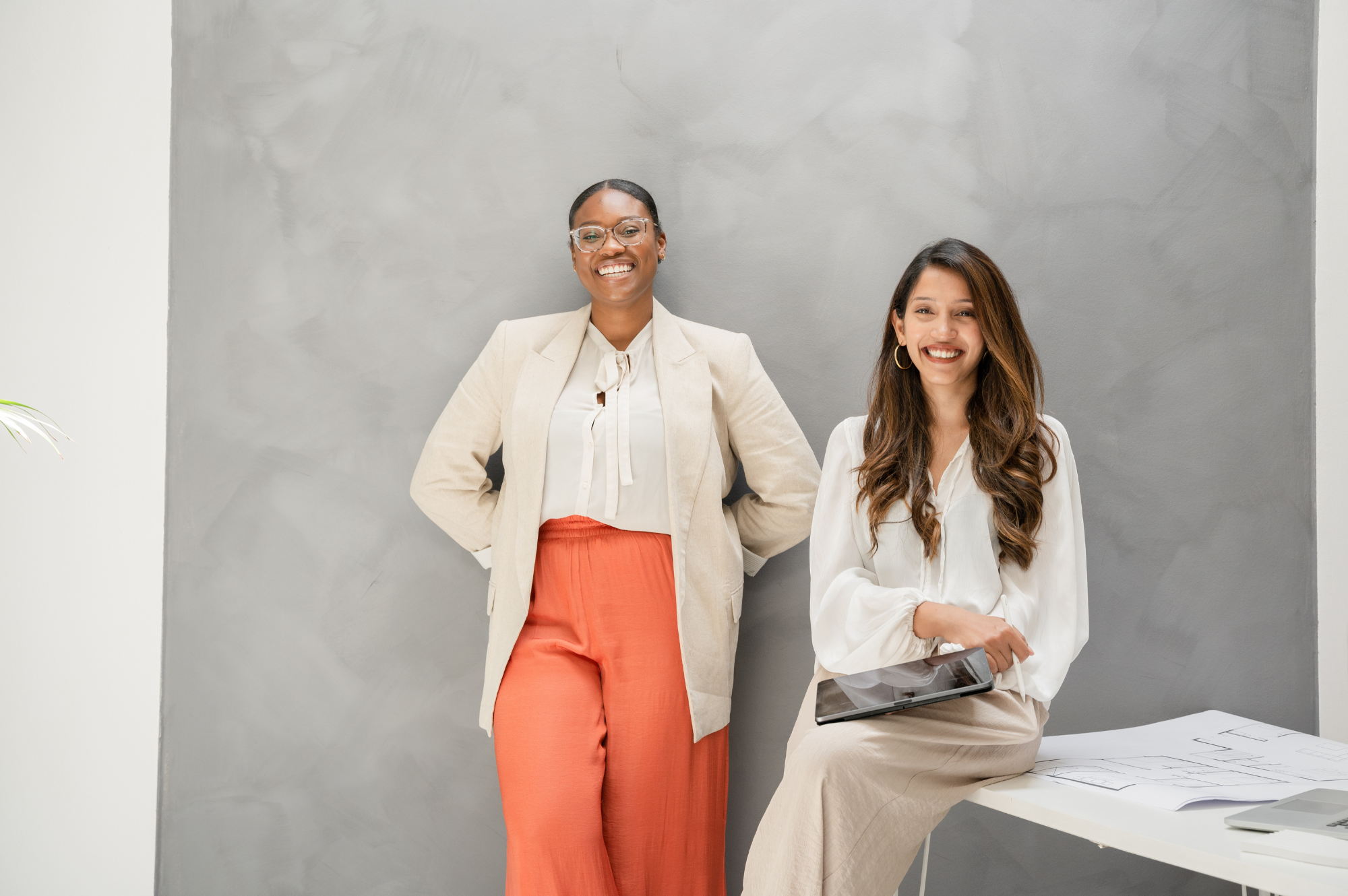 Two women smiling indoors, one standing and one sitting with papers and a tablet, against a gray wall.