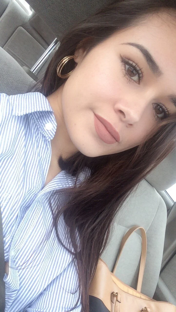 A young woman with brown hair wearing hoop earrings and a blue and white striped shirt, sitting in a car. There is a tan purse on the seat beside her.