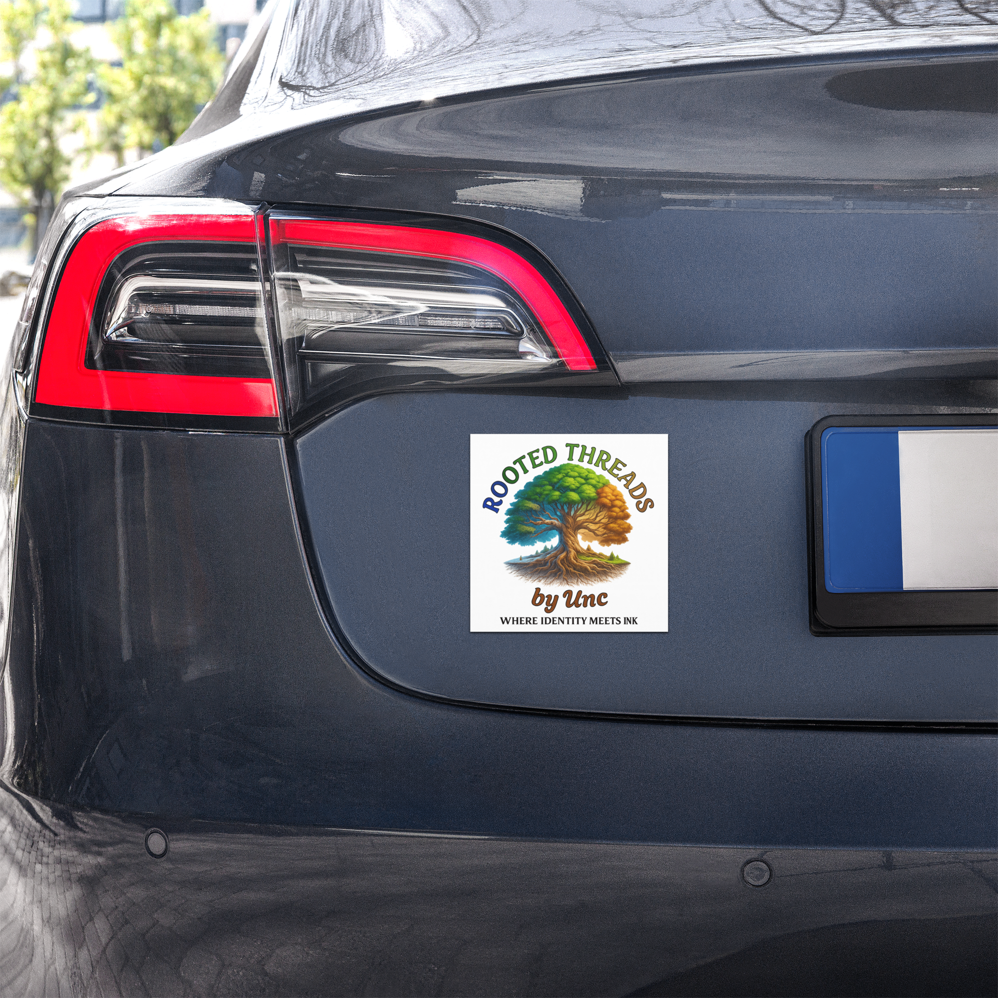Close-up of the rear left side of a dark-colored car, showing part of the taillight and a sticker with a tree and the text 'RootedThreads by Unc'.