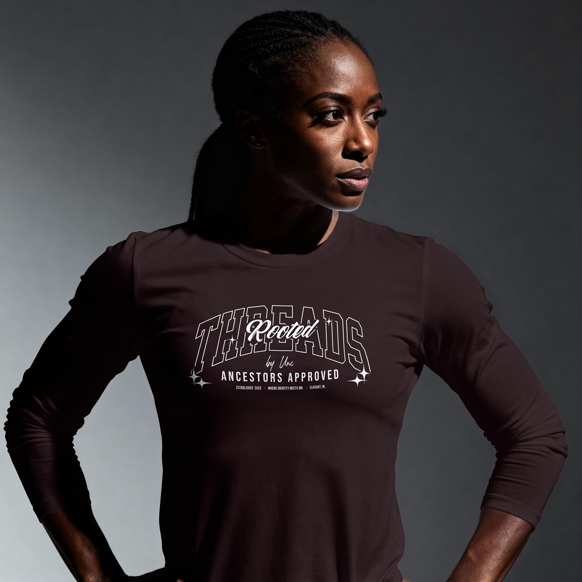 A woman with dark skin and braided hair looking to the side, wearing a dark long-sleeve shirt with white text that reads 'Rooted Threads by Unc' and smaller text underneath that says 'Ancestors Approved'.