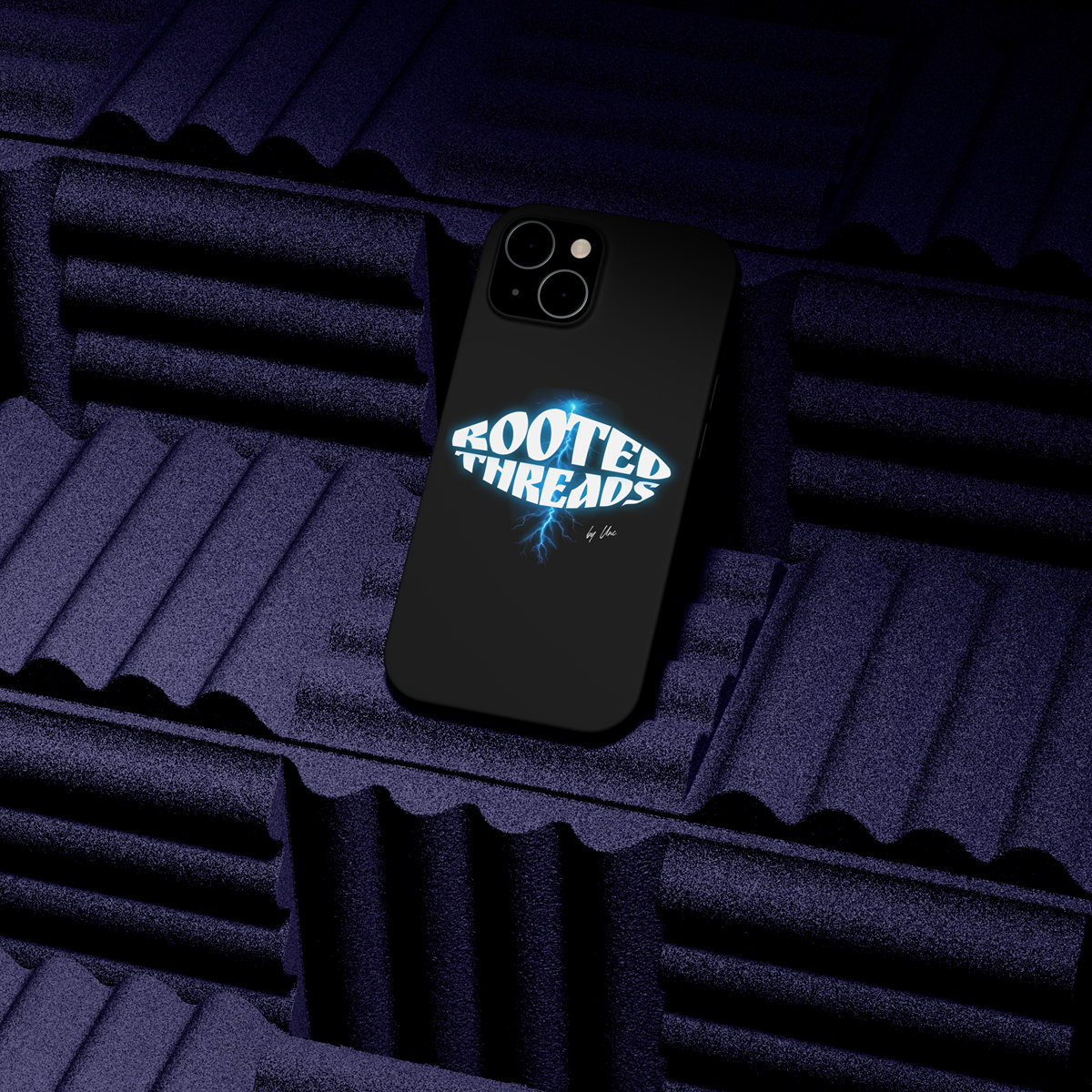 A black phone case with the logo 'Rooted Threads' in bold, glowing blue and white letters, featuring a lightning bolt underneath. The phone is placed on purple soundproof foam panels.