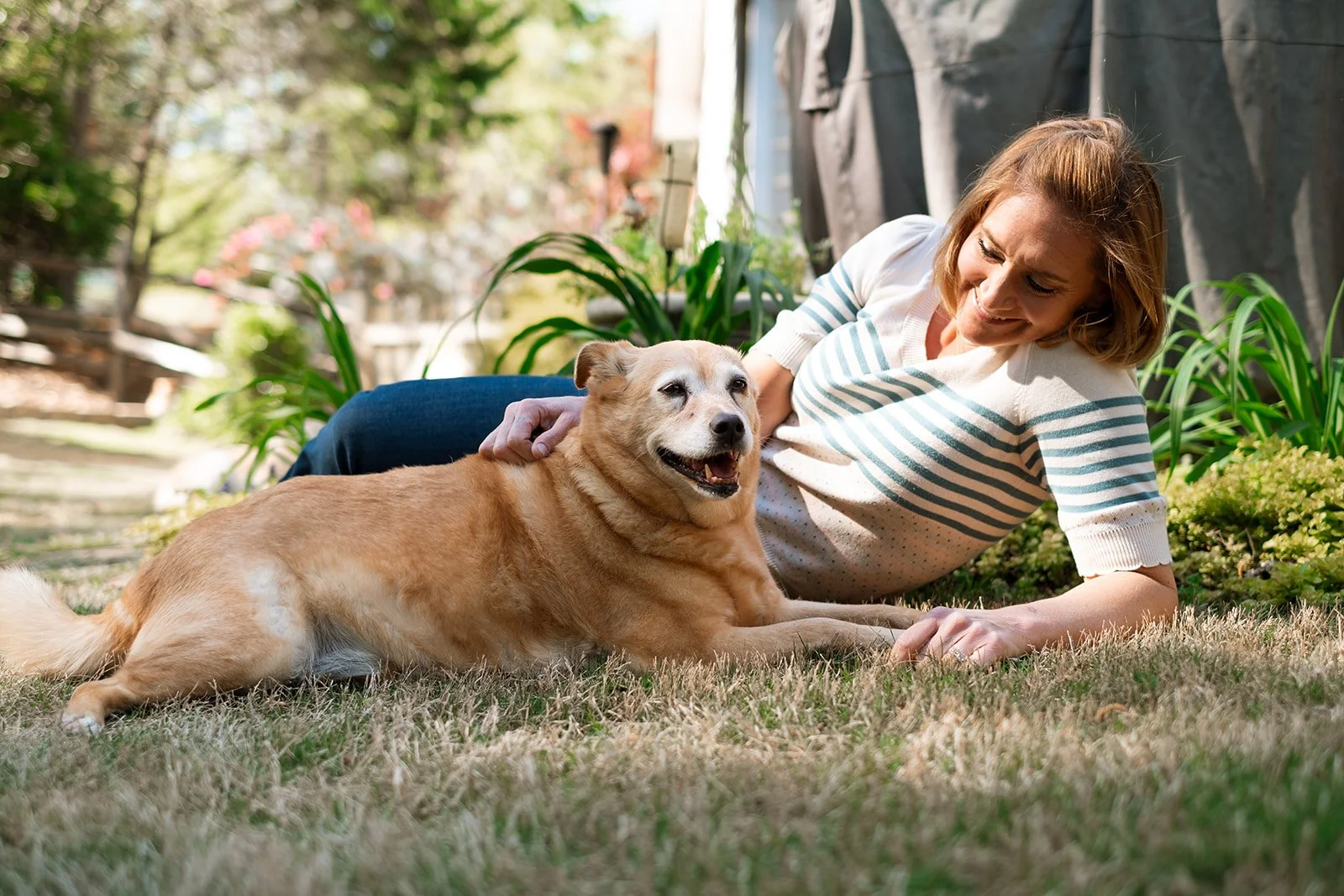 A woman lying on the grass outdoors, petting a golden retriever dog. Trees and plants are visible in the background on a sunny day.