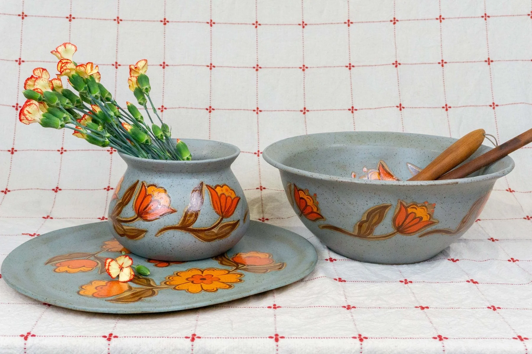 Ceramic vase with painted flowers containing yellow and orange carnations, ceramic plate and ceramic bowl with matching painted flower designs, set on a white cloth with red grid pattern.