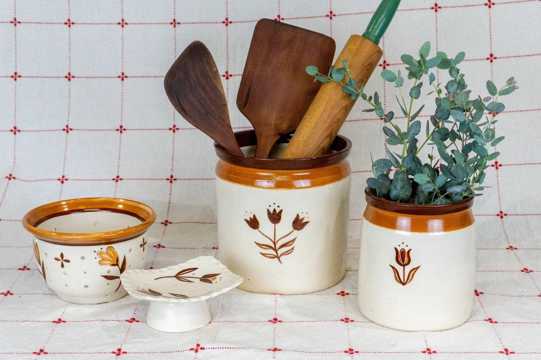 Set of rustic cream-colored kitchenware with brown decorative accents, including jars, a small bowl, and a plate, with wooden utensils and a small green potted plant against a white and red checked background.
