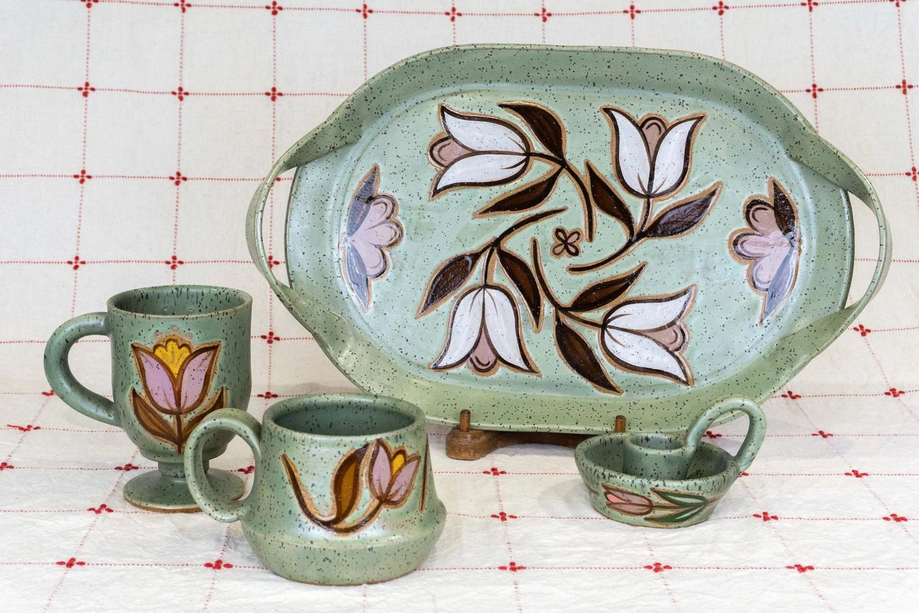 Set of green ceramic dishware with floral and tulip patterns, including a large oval tray, two coffee mugs, and a small bowl, displayed on a white cloth with red grid pattern.