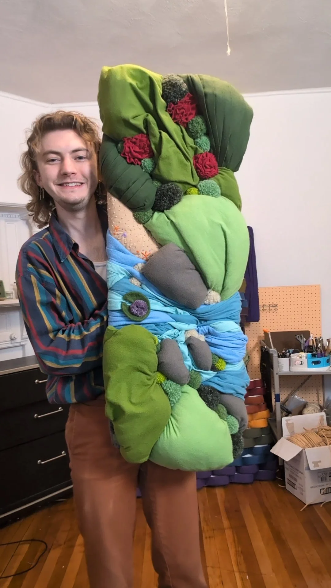The artist, Jacob Keven, holds their work, topography, in their studio. The work is a landscape made of textiles with colorful green hills and a blue river rushing over rocks next to a sandy beach.