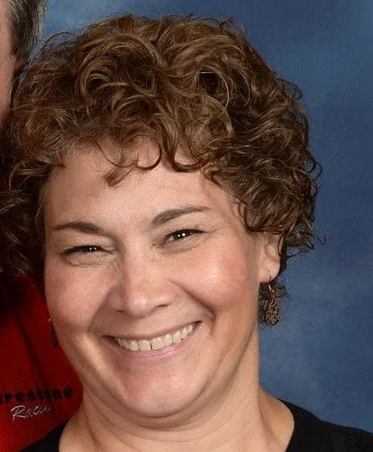 Close-up of a smiling woman with short, curly brown hair and earrings, against a blue background.
