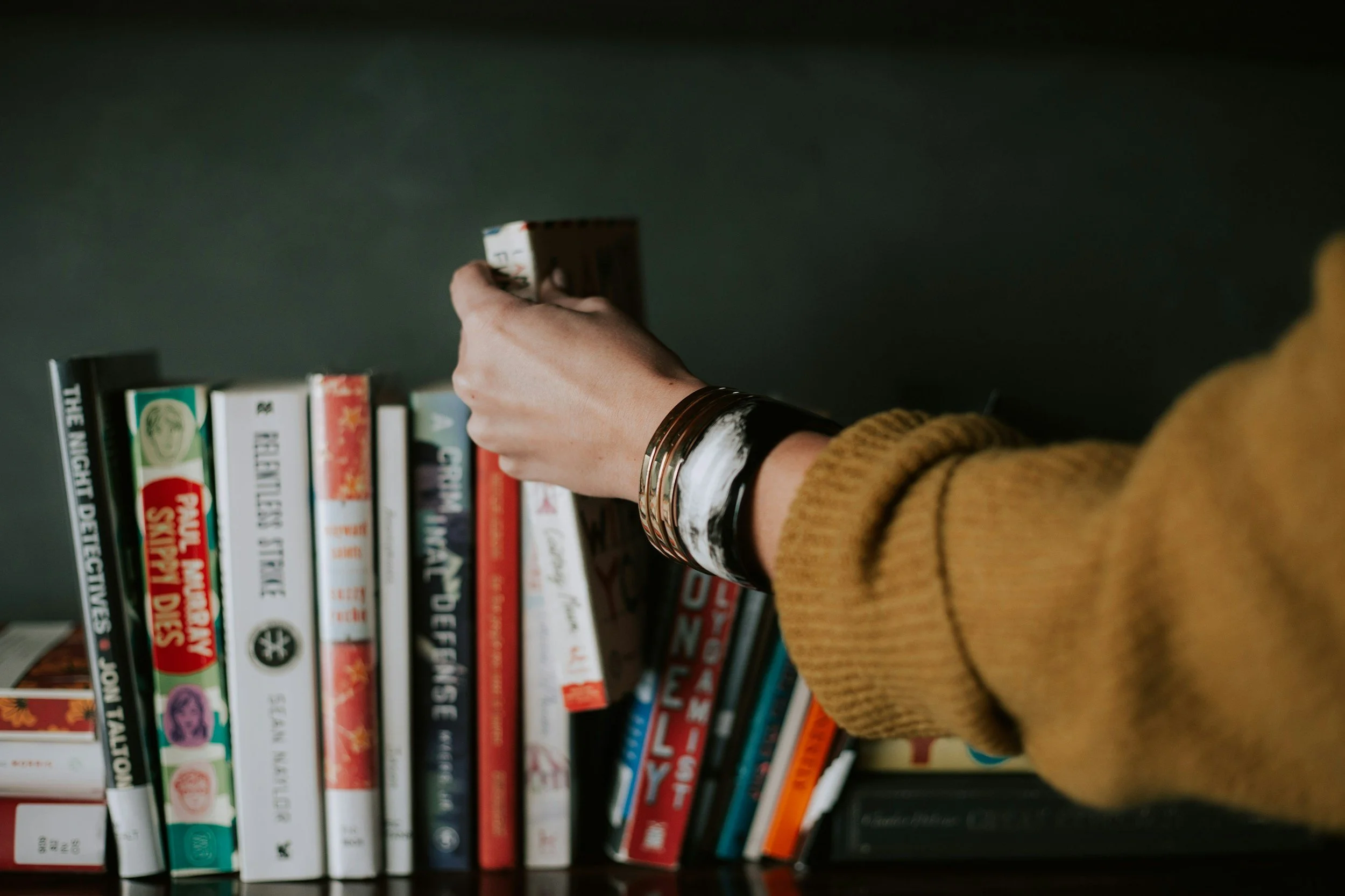 Person reaching for a book on a bookshelf with a green wall, wearing a mustard-colored sweater and gold bracelets.