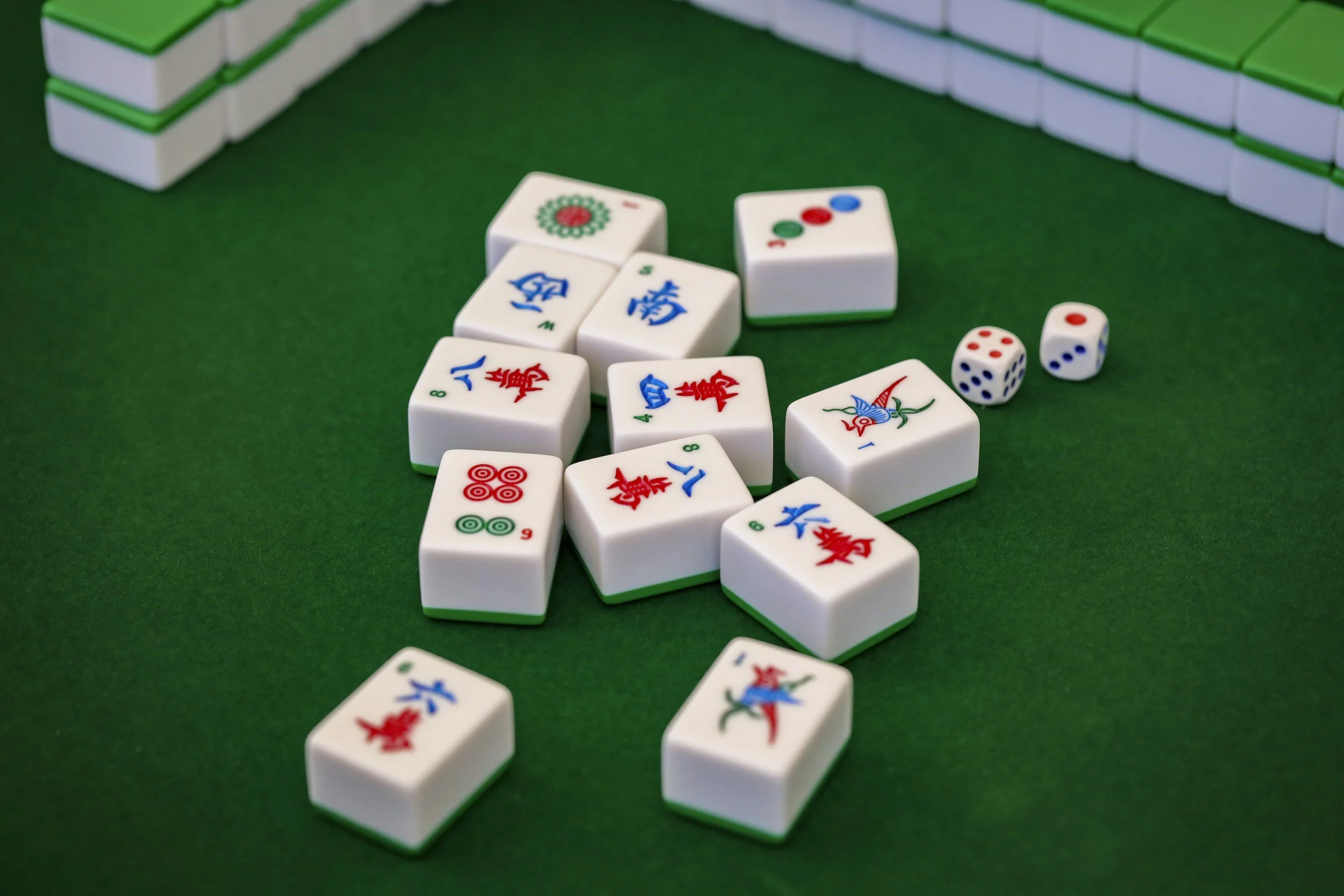 A Mahjong game setup with white tiles decorated with colorful symbols, dominoes, and two white dice with red and blue dots on a green felt surface. In the background, there are stacks of Mahjong tiles.