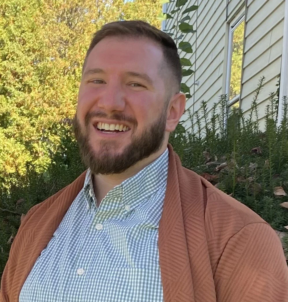 A young man with a beard smiling outdoors, wearing a checked shirt and a brown jacket, standing in front of bushes and a house with siding.