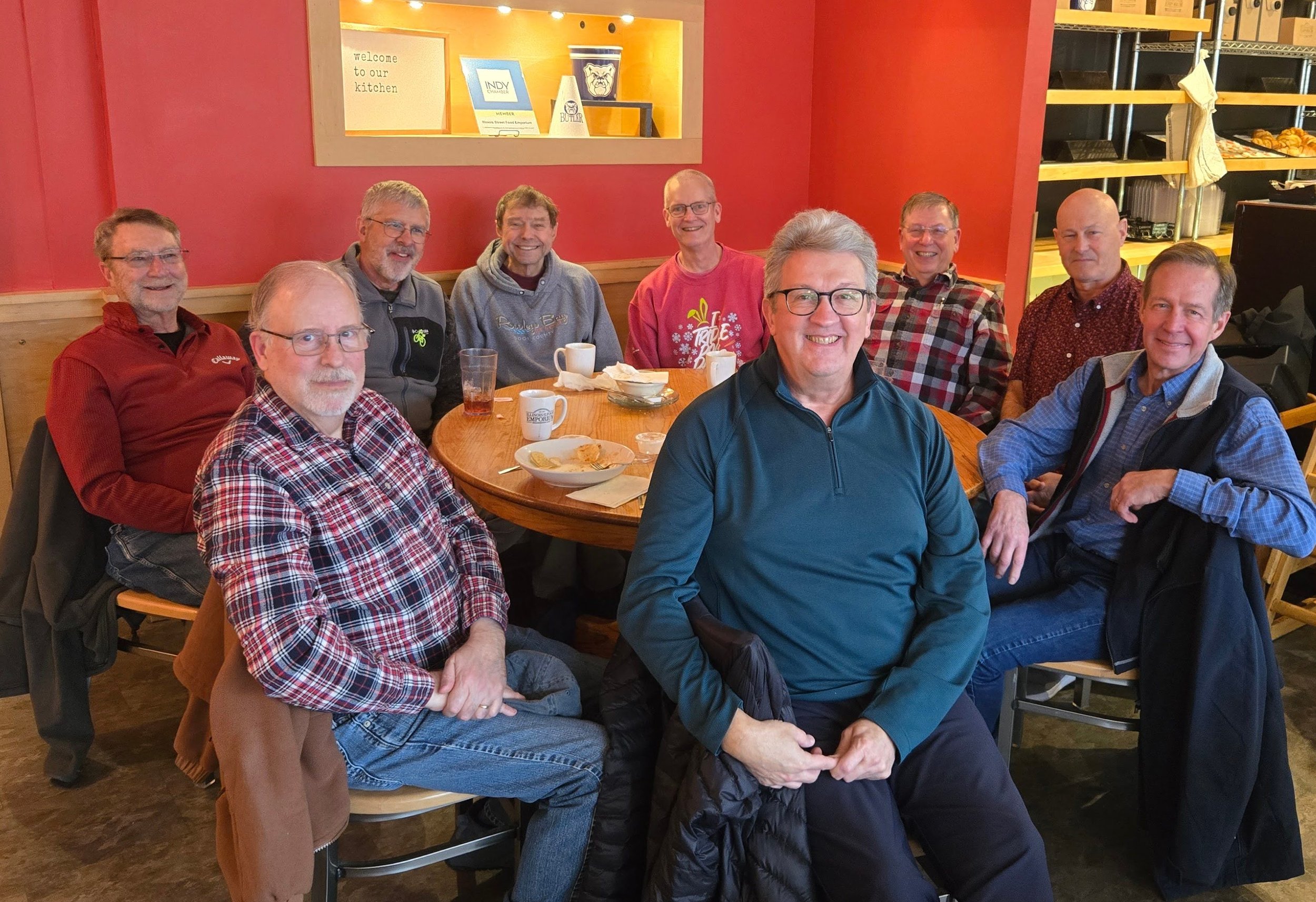 A group of nine men sitting around a wooden table in a restaurant, smiling at the camera. The restaurant has red walls, and there are plates, cups, and food remnants on the table.