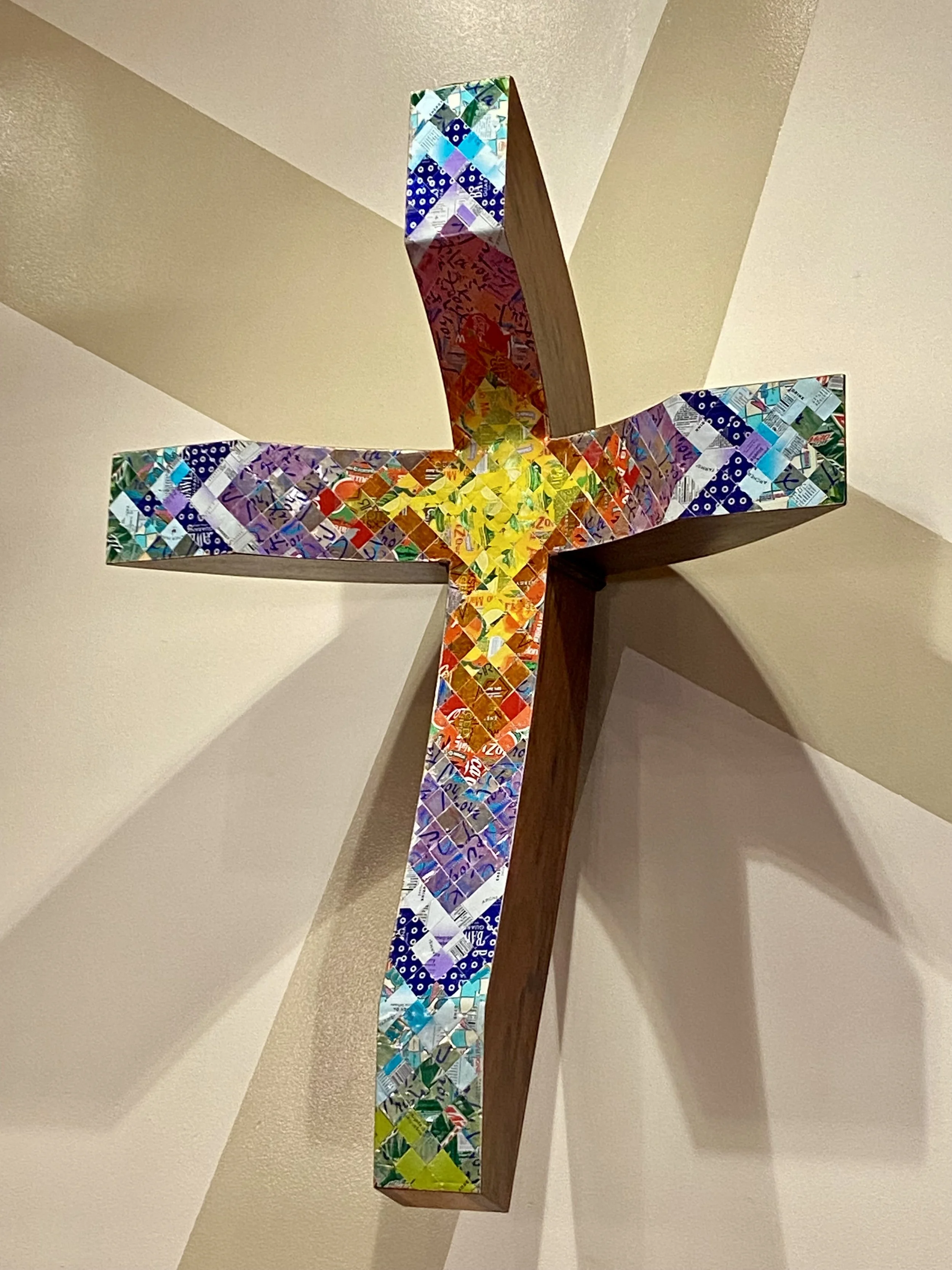A wooden cross with colorful mosaic inlays featuring various patterns, mounted on a beige wall.