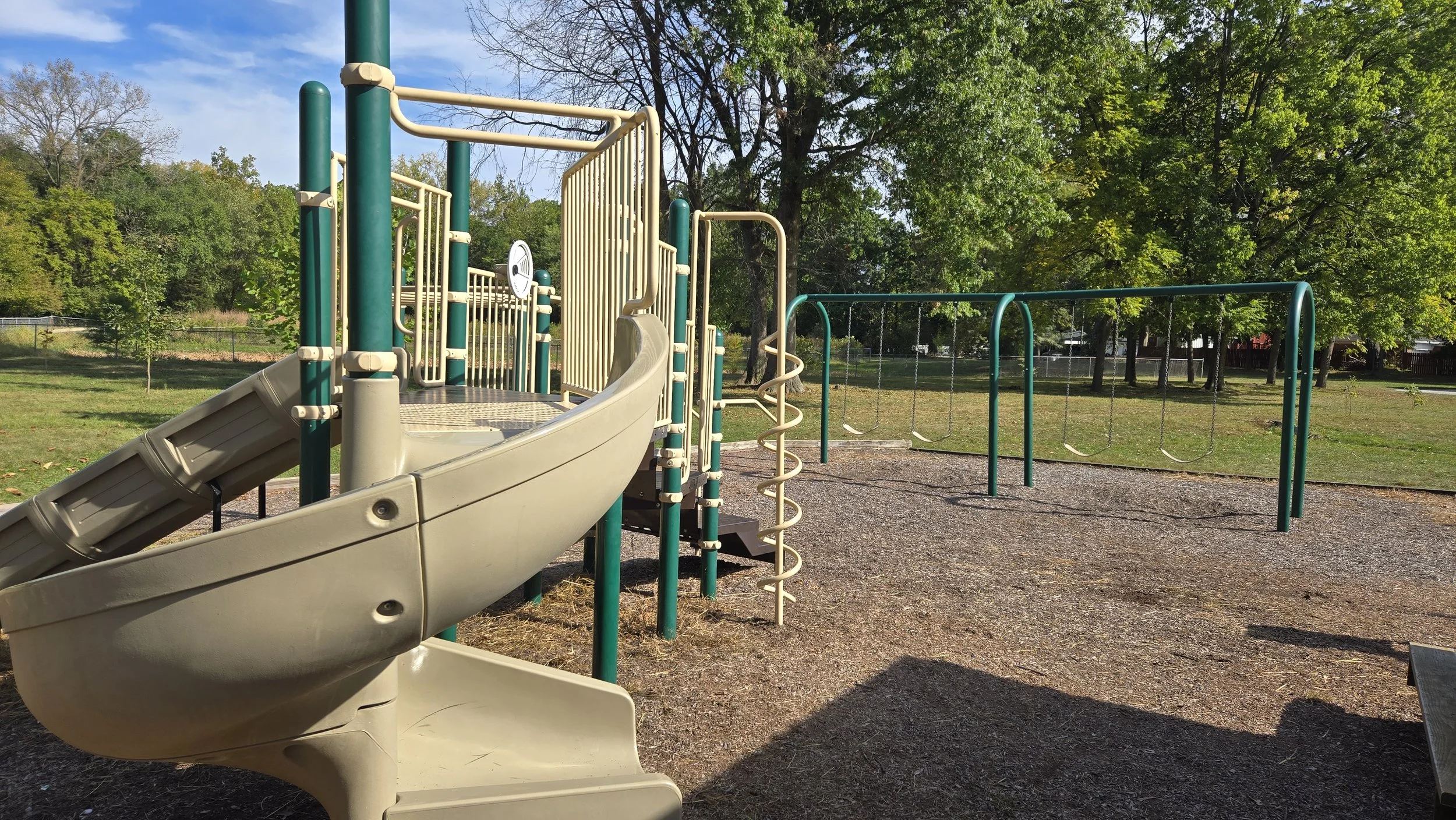 Children's playground with a slide, climbing structures, and swings in a park with green trees and some open grassy areas.