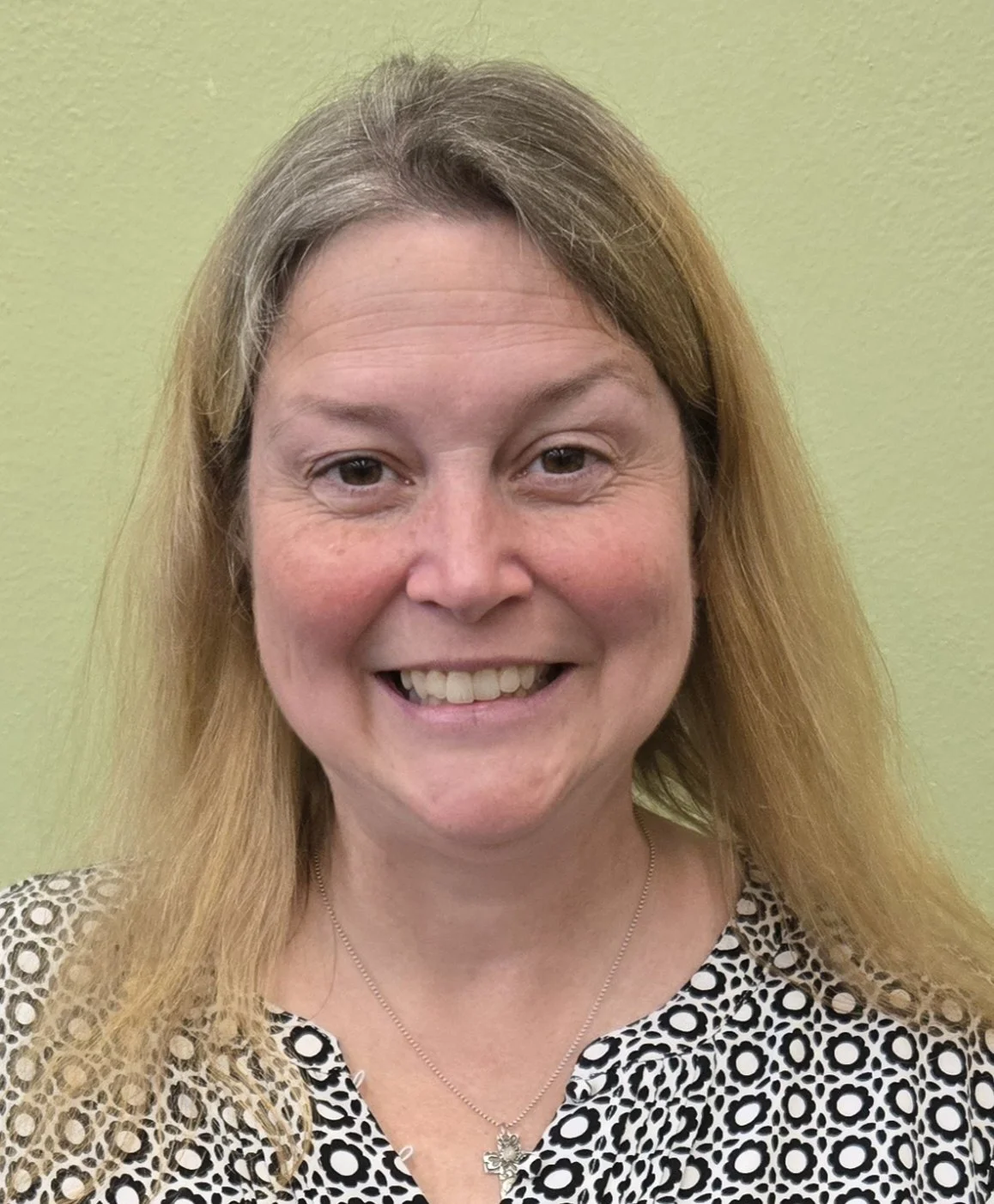 A woman with light brown hair and light skin smiling in front of a plain light green wall, wearing a black and white patterned blouse and a necklace with a cross pendant.