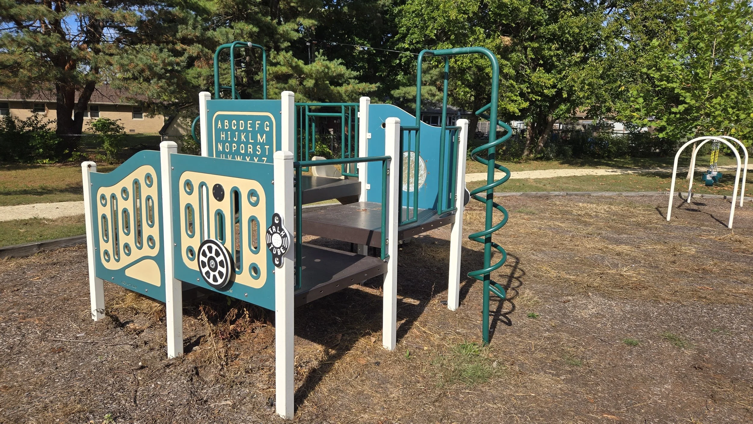 Colorful playground equipment featuring a small climbing structure with blue and white panels, a black slide, a green spiral climber, and a swing set in the background, set in a park with trees and a walking path.