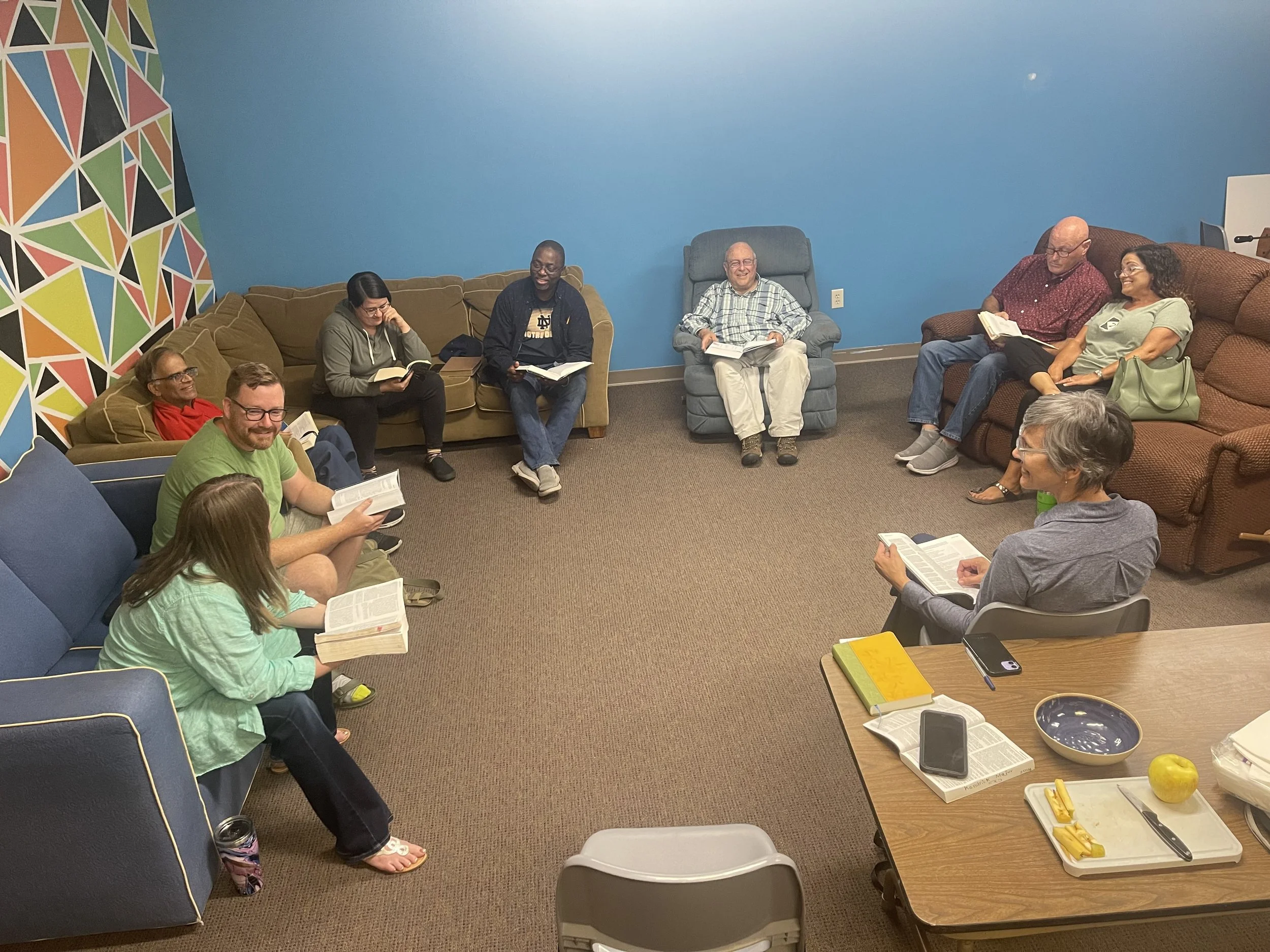 A group of ten people sitting in a circle on couches and chairs, reading from books, in a room with blue walls and a colorful geometric mural on one wall.