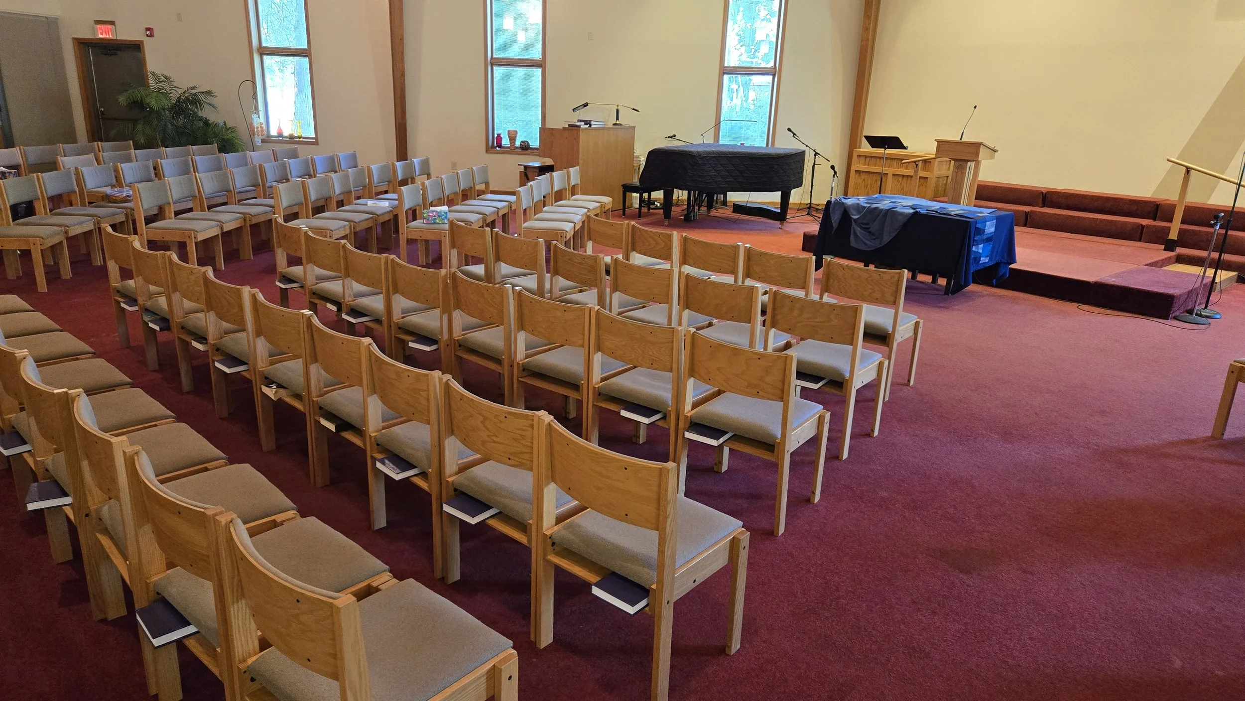 An empty church or auditorium with wooden chairs arranged in rows facing a stage. The stage has a grand piano, a pulpit, and microphones, with windows on the back wall letting in natural light. The room has a red carpet and wooden accents.