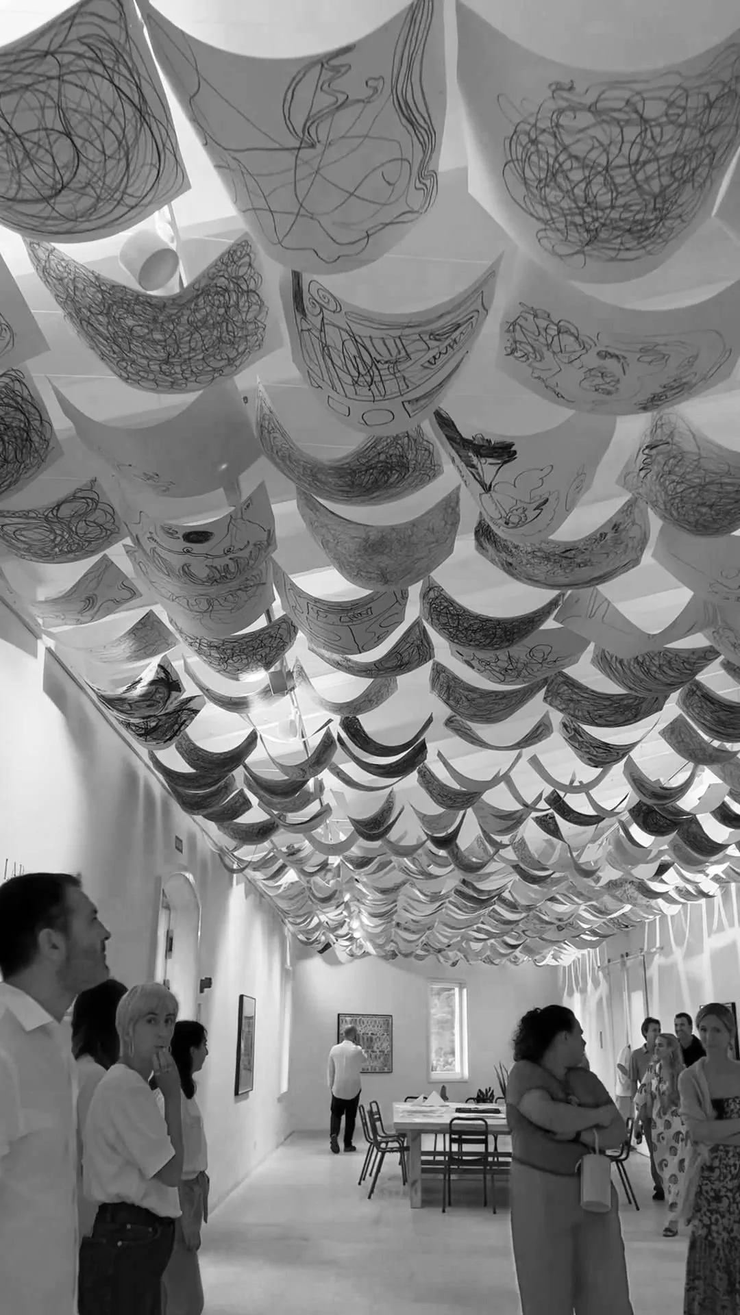 An art gallery with numerous sketches hanging from the ceiling. People are observing and discussing the artwork, with tables and chairs visible in the background.