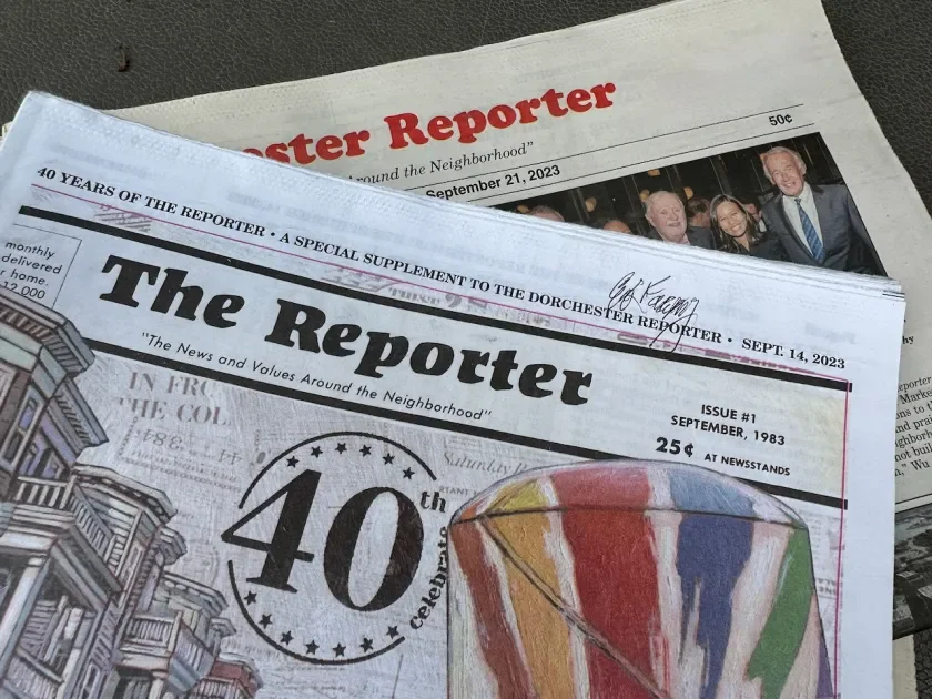 A man with curly dark hair, glasses, and a beard looking to the right, wearing a black sweater and sitting indoors with a plant and picture frame in the background.