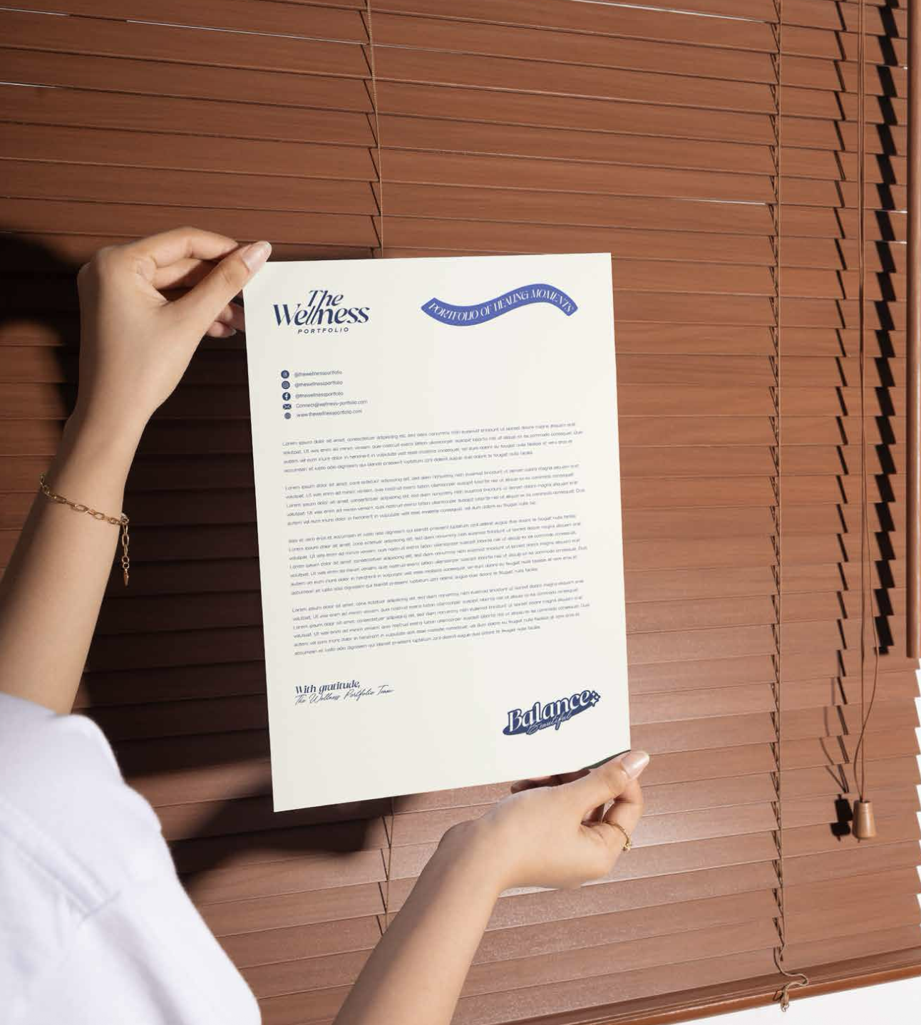 Person holding a printed letter or portfolio titled 'The Wellness Portfolio' with social media icons and contact information, against brown wooden blinds.