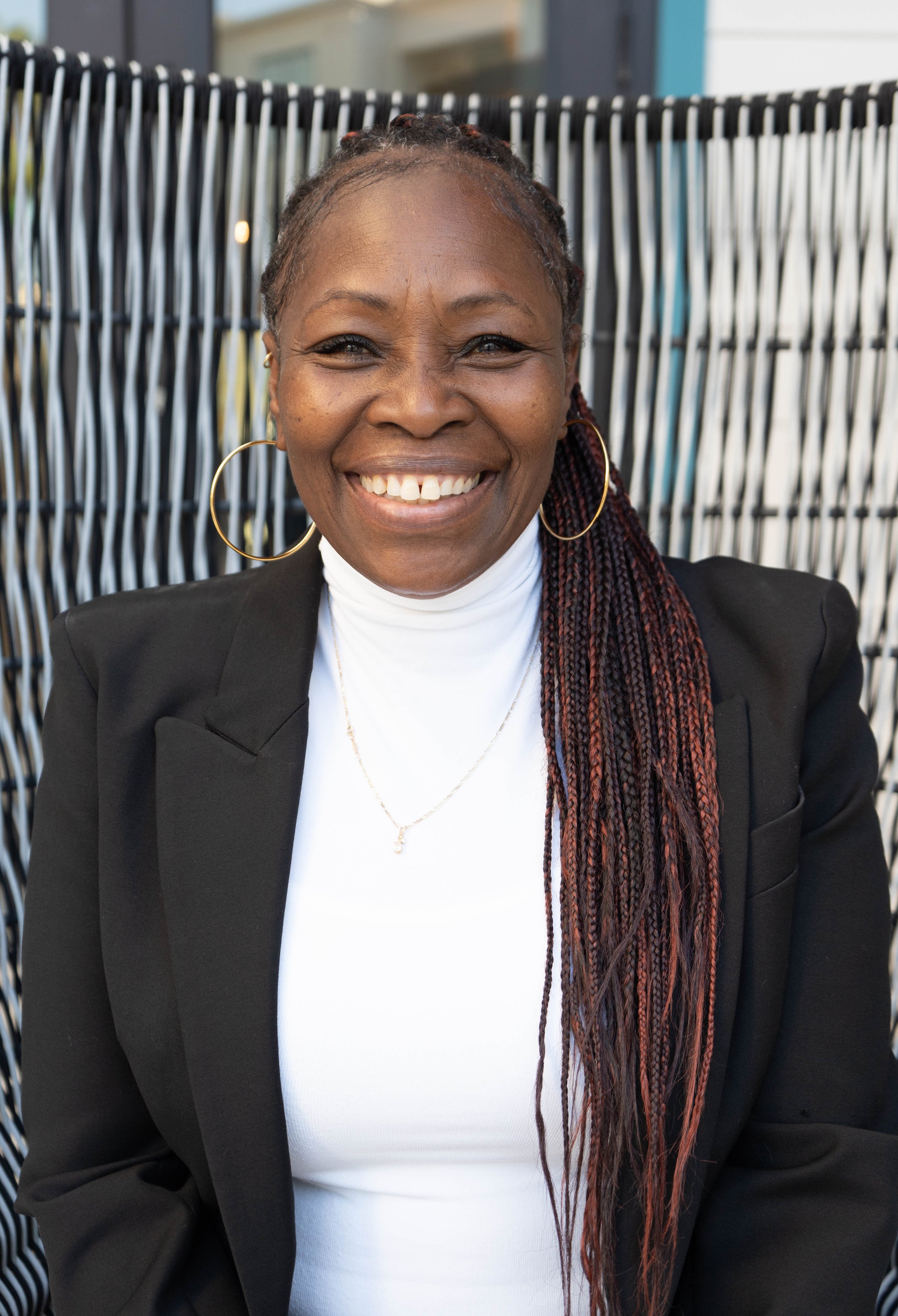 A smiling woman with long braided hair, gold hoop earrings, wearing a white turtleneck and black blazer, seated in front of a patterned outdoor background.