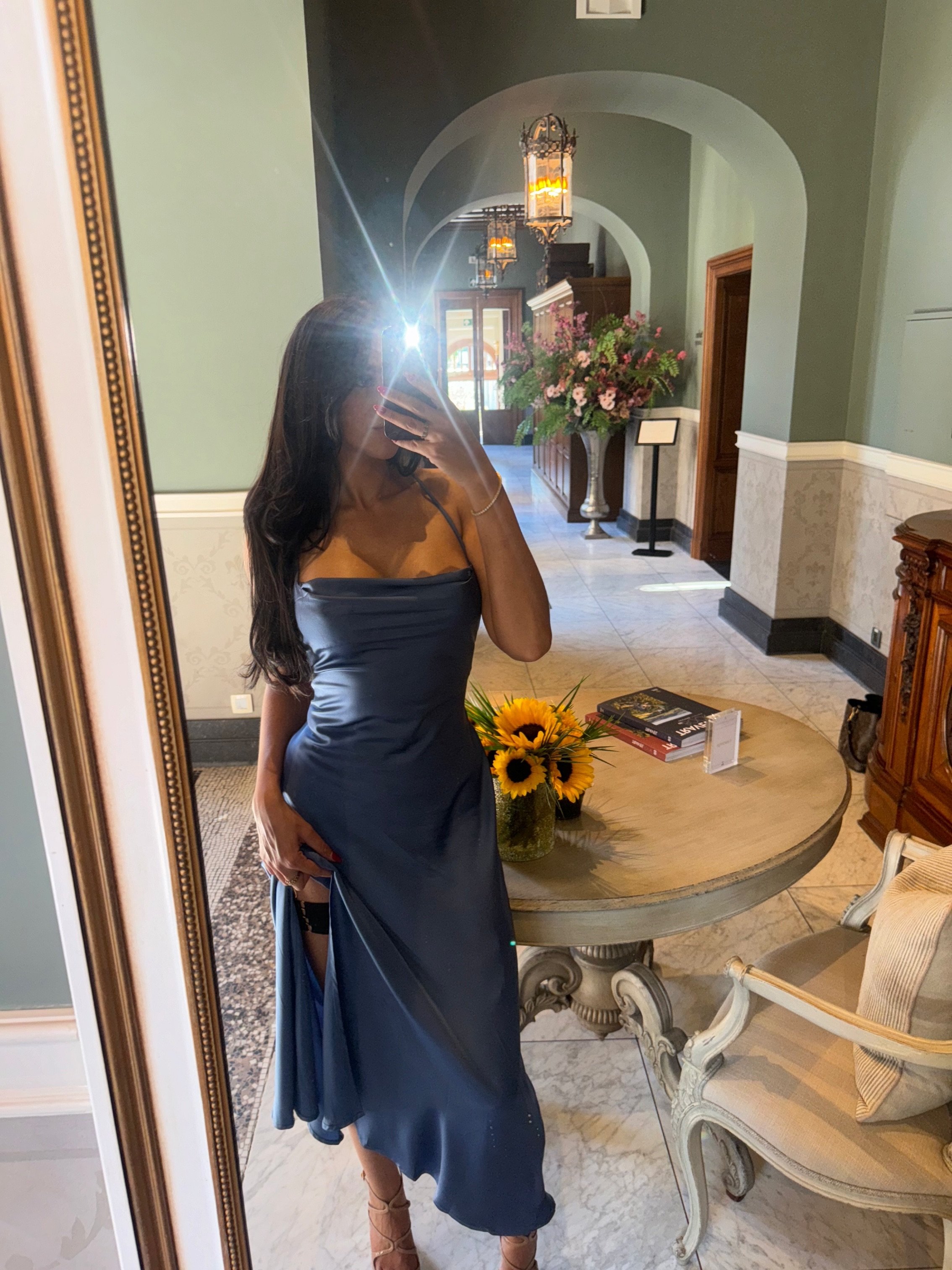 A woman with long dark hair taking a mirror selfie with her phone, wearing a dark blue satin dress, in a well-decorated lobby or reception area with flowers, books, and elegant furniture.
