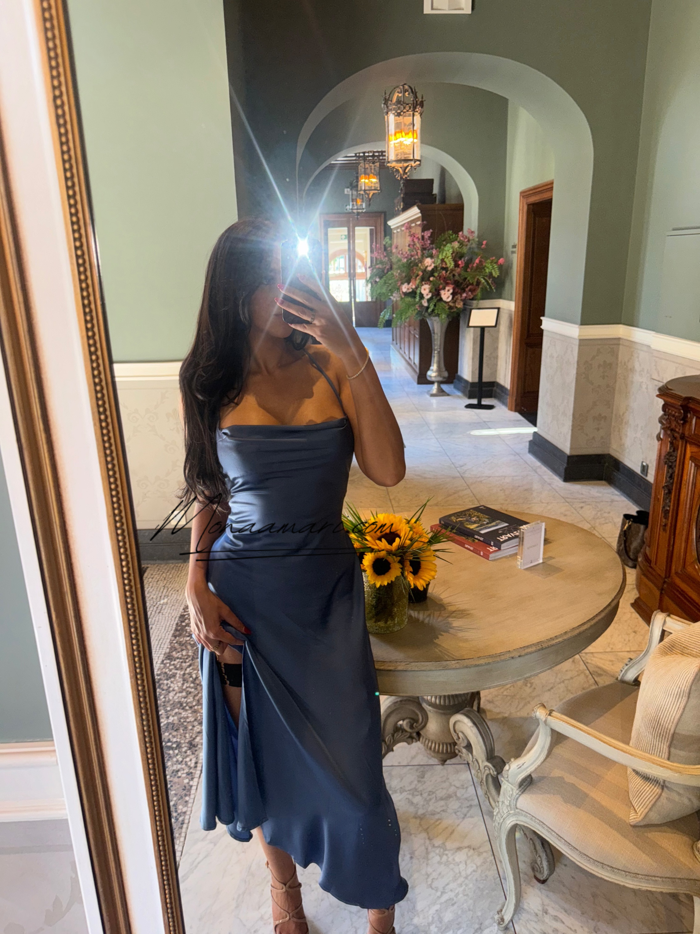 Woman taking a mirror selfie, wearing a navy blue satin dress with thin straps, in a hotel lobby or reception area with a round wooden table, sunflower vase, a bouquet of flowers, books, and cushioned chairs.