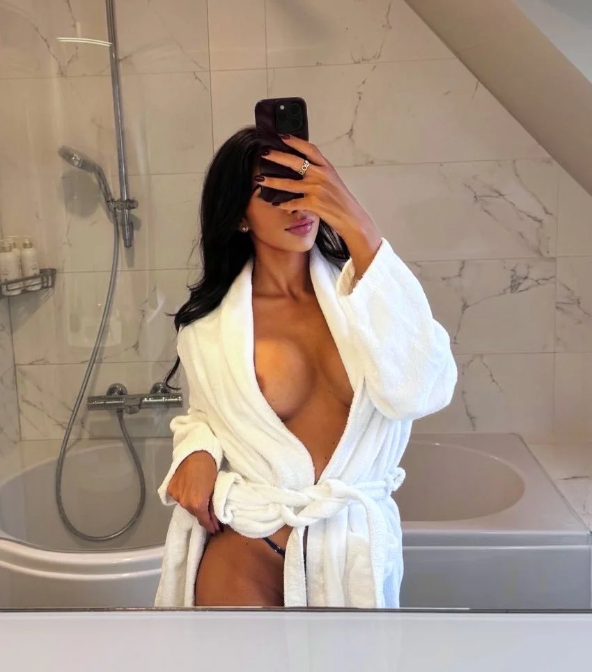 A woman in a white bathrobe taking a mirror selfie in a bathroom, with her face partially covered by her phone.