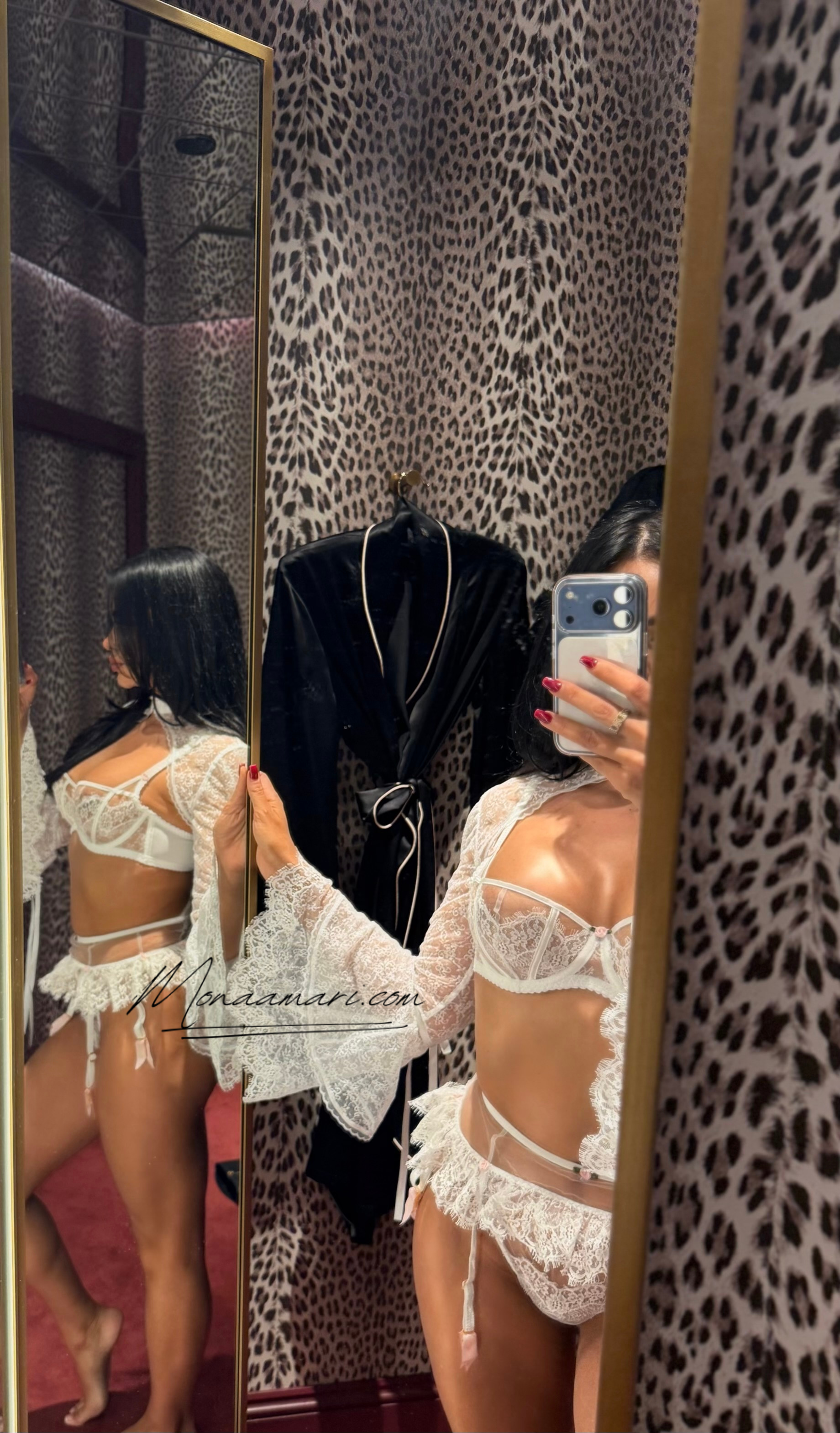 A woman taking a mirror selfie in a dressing room with leopard print walls, wearing white lace lingerie, and holding a smartphone. There is a black robe hanging on the wall.