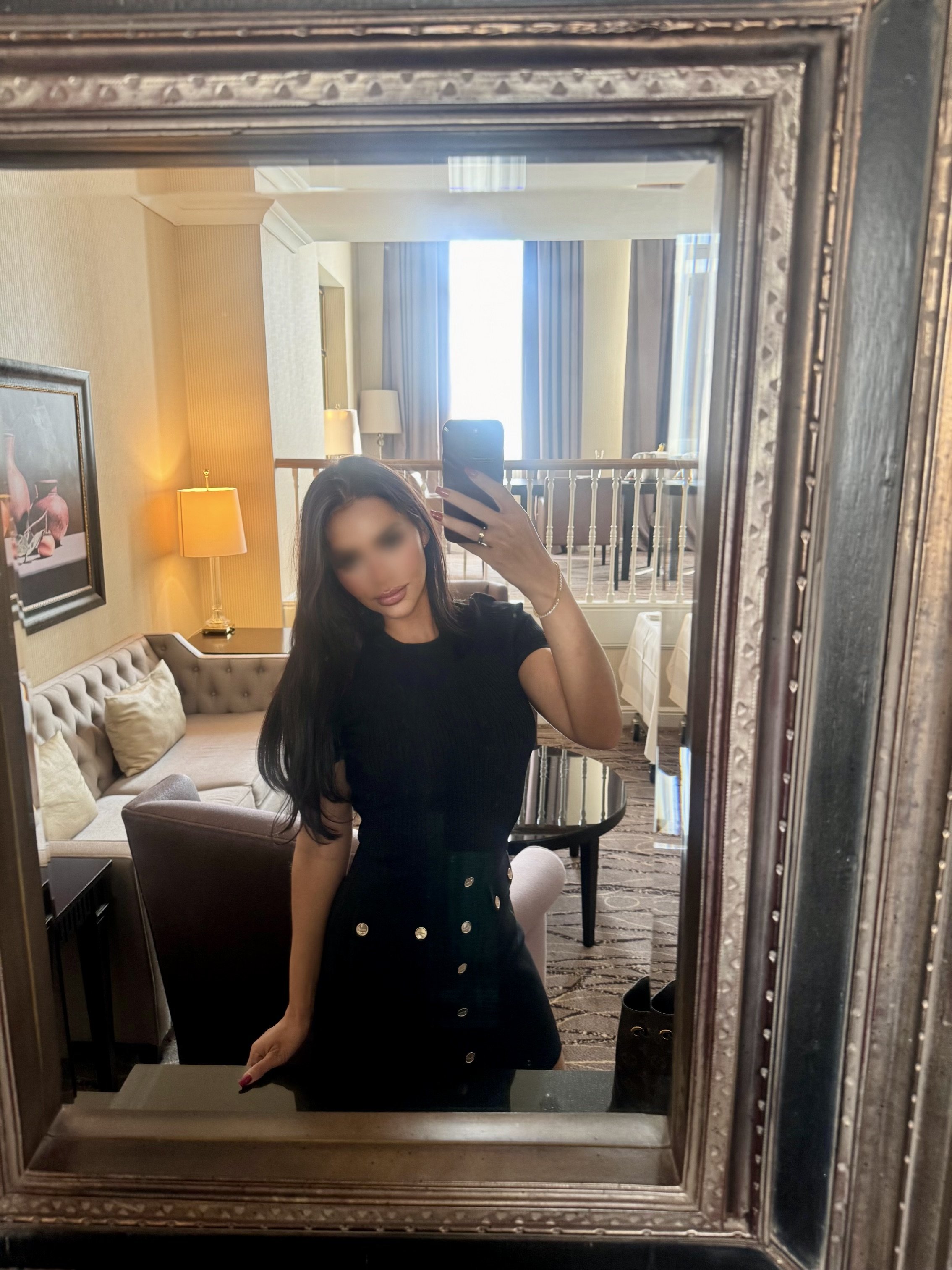 A woman taking a mirror selfie in a hotel room, with a beige sofa, artwork, lamps, curtains, and a window in the background.