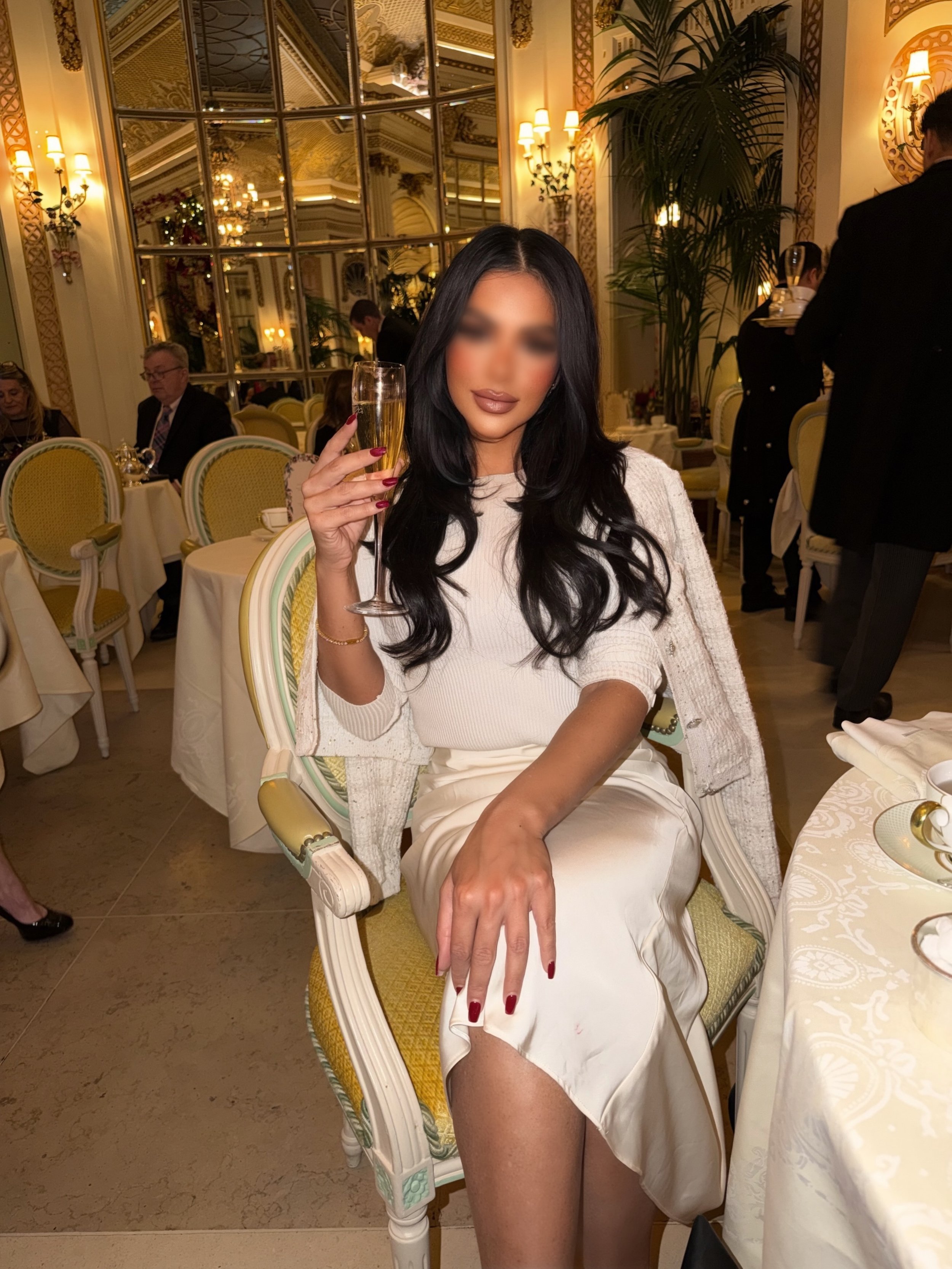 A woman with long black hair and red lipstick sitting on a green and white ornate chair in a fancy, well-lit restaurant. She is holding a champagne flute and wearing a white dress with a matching cardigan draped over her shoulders. The restaurant has elegant decor, large mirror panels, and other guests dining in the background.