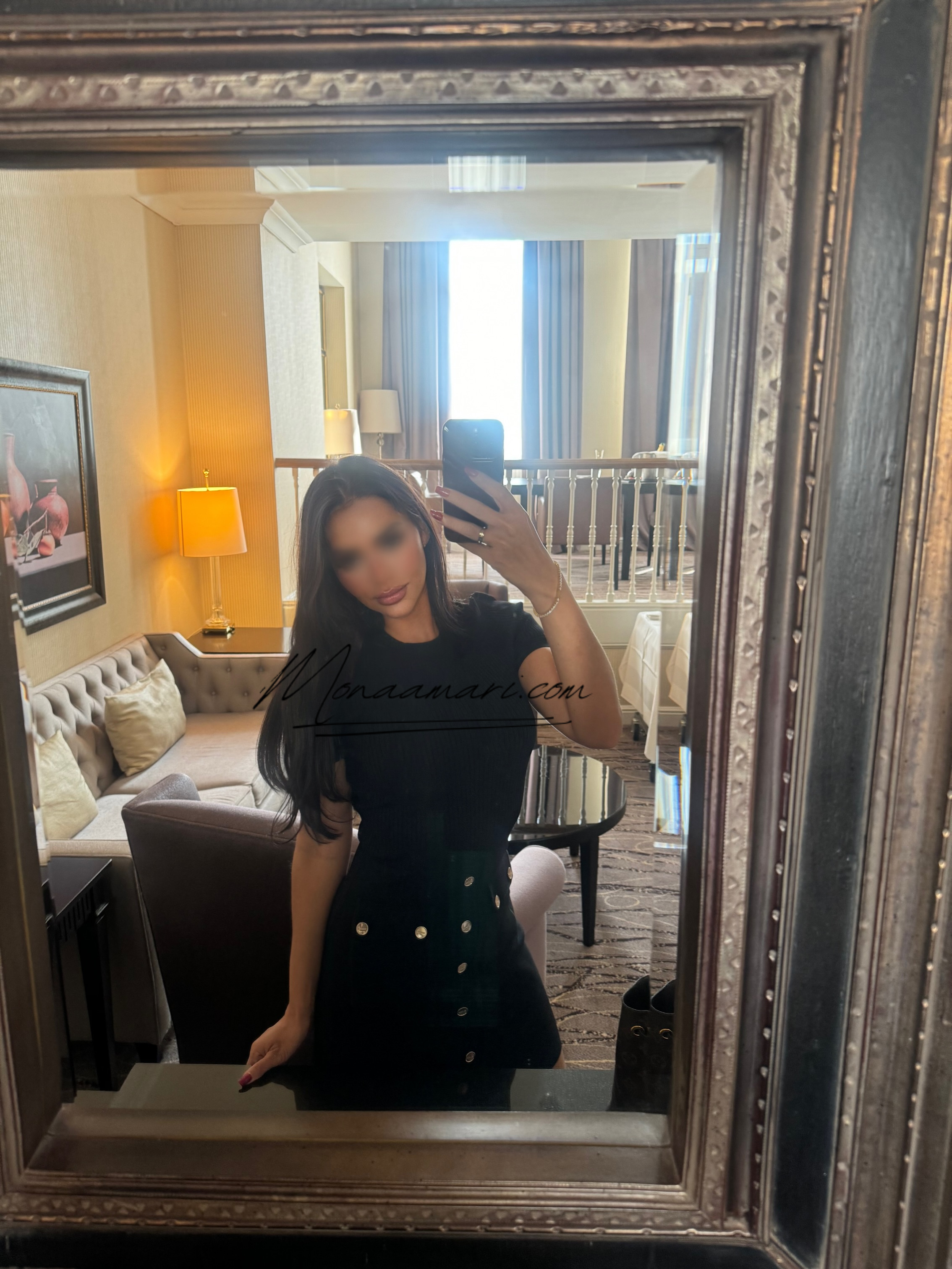A woman taking a mirror selfie in a hotel room, wearing a black dress with silver buttons.