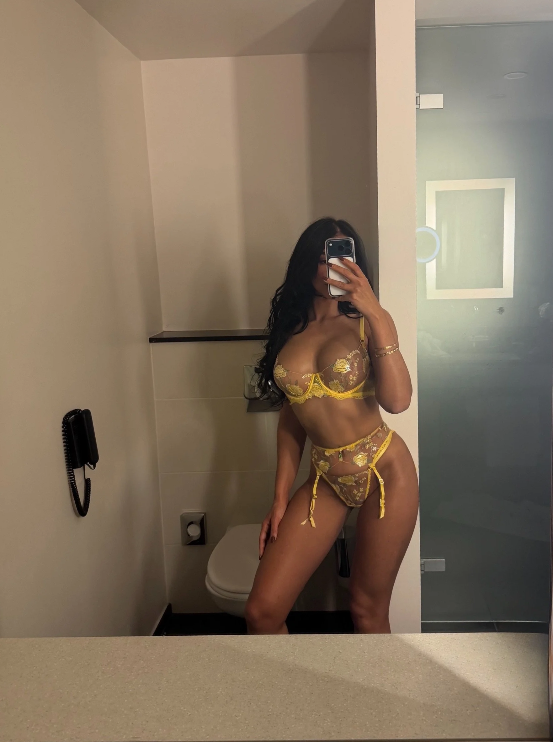 A woman taking a mirror selfie in a bathroom, wearing yellow floral lingerie with transparent details and matching garter belt, with long dark hair and gold bracelets.
