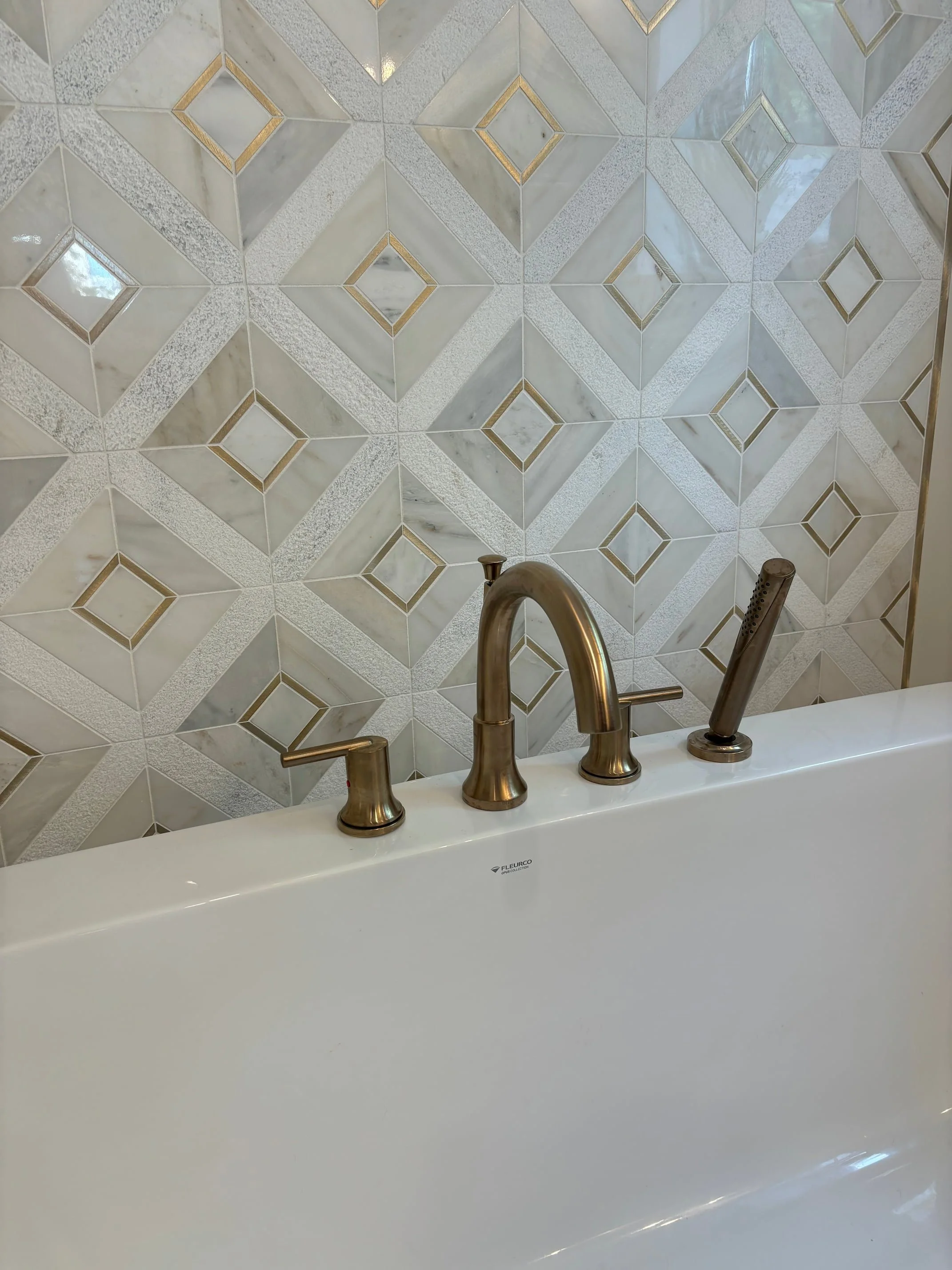Close-up of a white bathtub with brushed nickel faucet and handheld showerhead, in front of a tiled wall with geometric pattern of marble and gold accents.