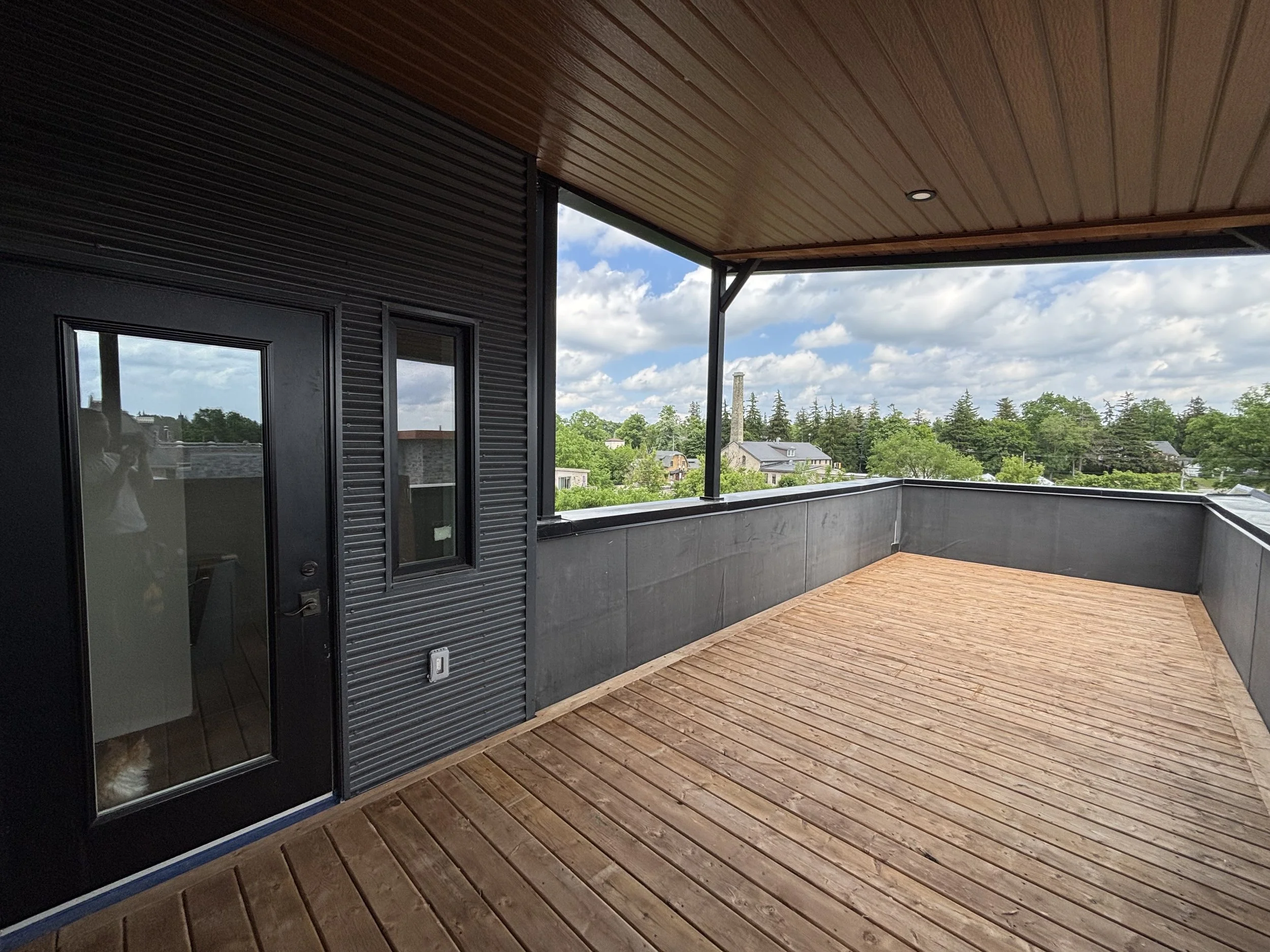 View of a balcony with wooden flooring, black metal walls, and a ceiling with wooden paneling, overlooking a neighborhood with trees and houses under a cloudy sky.