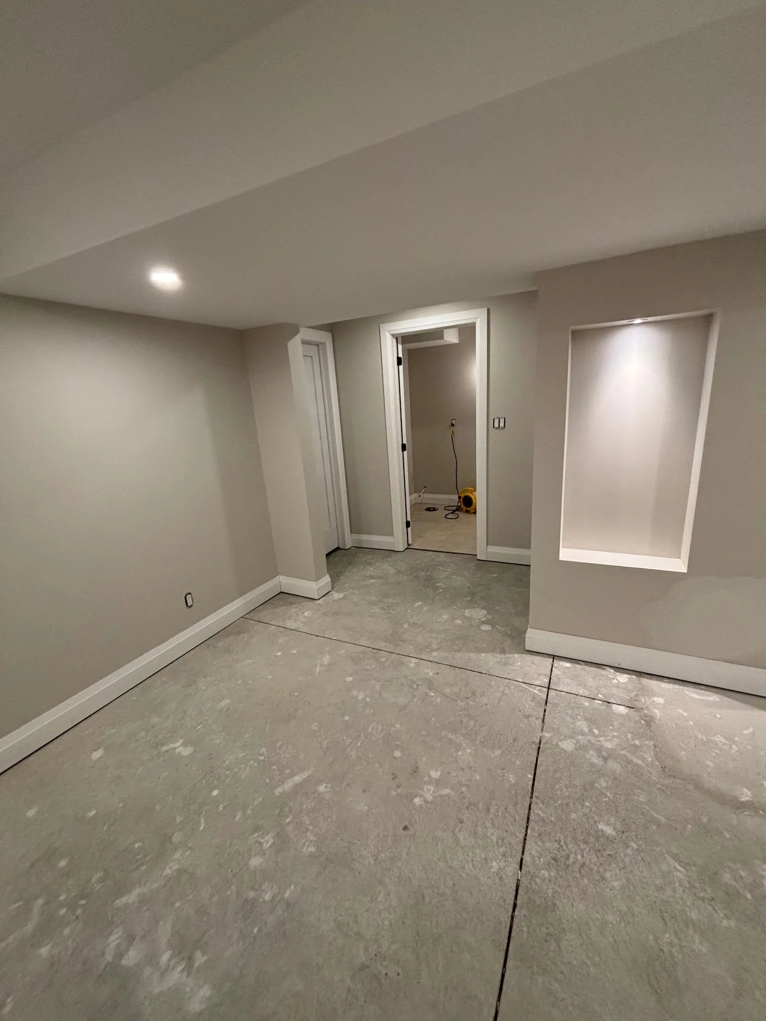 Interior view of a house under construction, showing unfinished floors with some debris, partially painted beige walls, with a doorway leading to another room where construction tools are visible.
