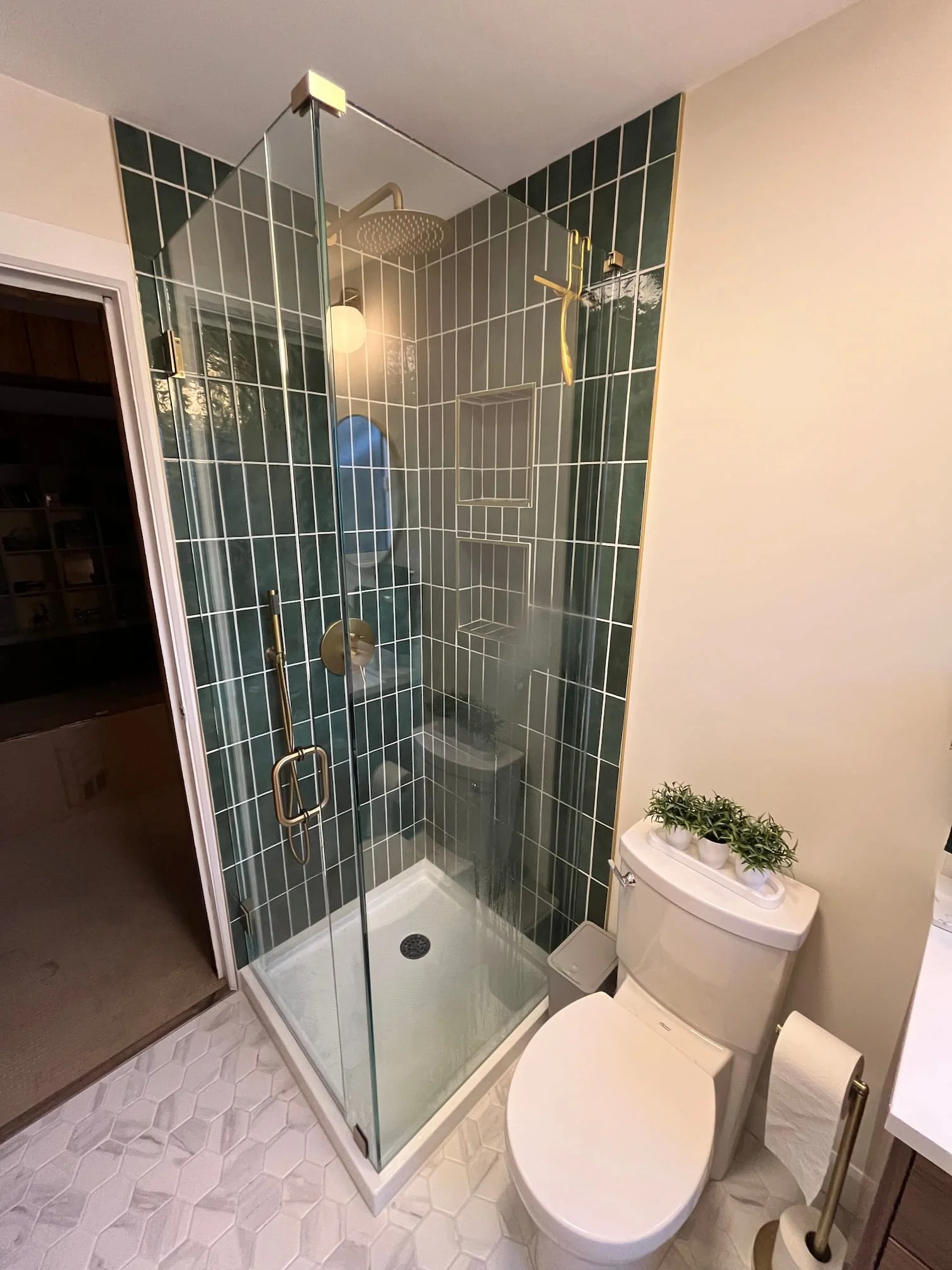 Small modern bathroom with a glass-enclosed shower, white hexagonal tile flooring, a toilet with a plant on top, and a white side table with a gold towel holder and beige wall.