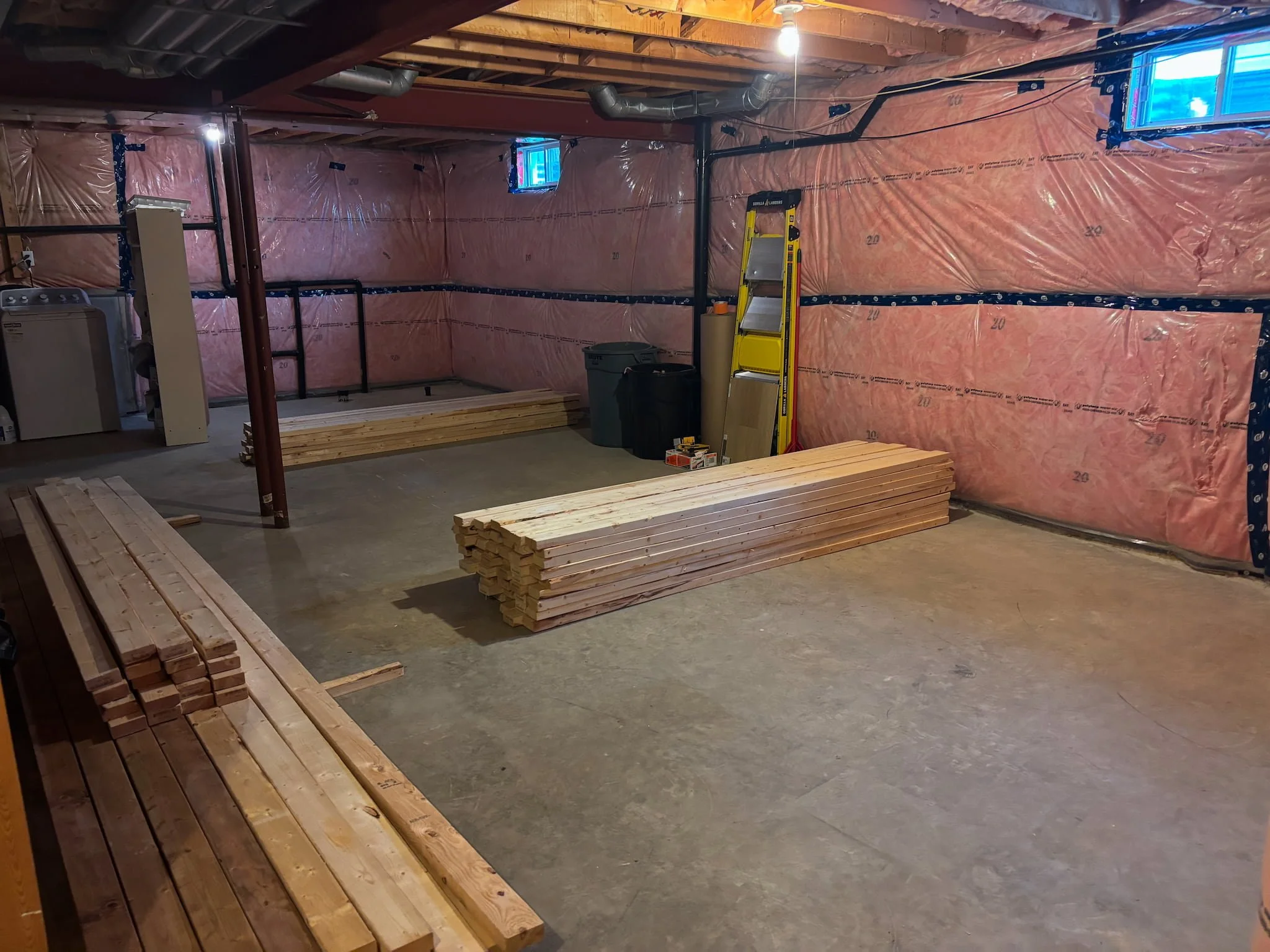 A basement construction area with exposed pink insulation on the walls, wooden beams, and a concrete floor. There are stacks of wooden planks and a yellow ladder. Small windows near the ceiling provide natural light, and there is a washing machine in