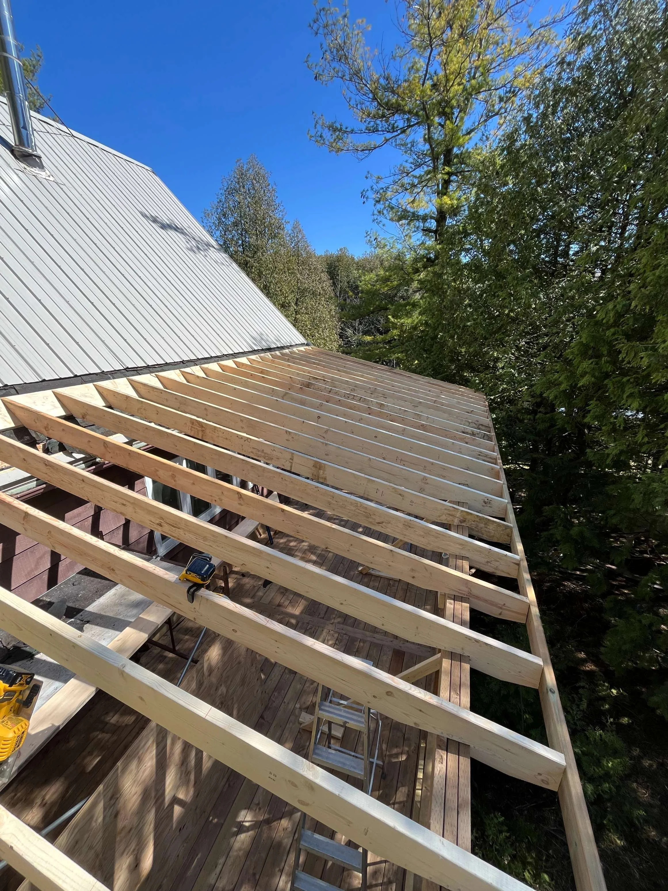 Construction site on a wooden deck in a forest, with a partially built roof frame, woodworking tools, and a metal roof in the background under a clear blue sky.