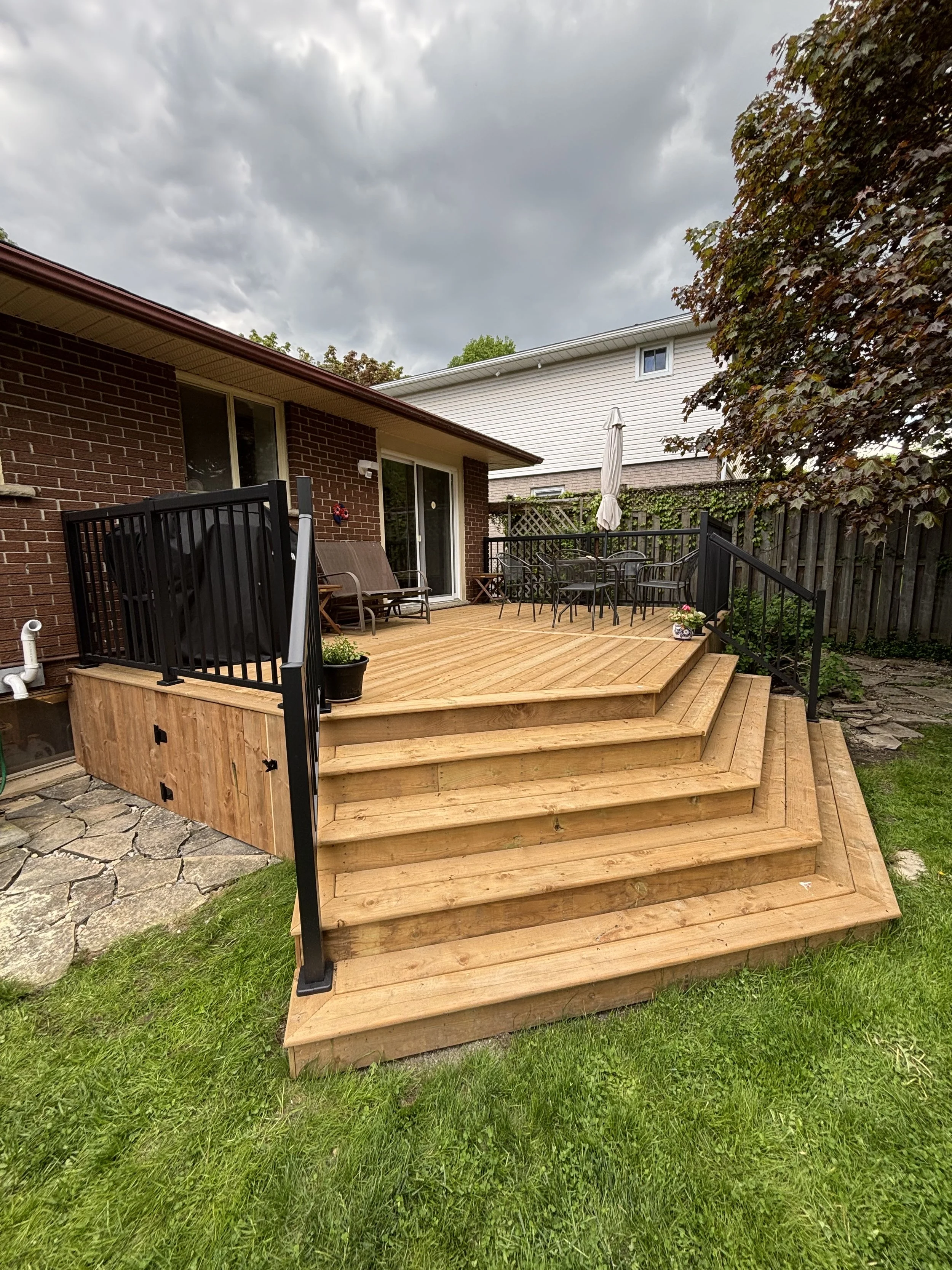 A wooden deck with stairs leading down to a grassy yard, outdoor furniture, and a screened porch area attached to a brick house. There are trees and an overcast sky in the background.