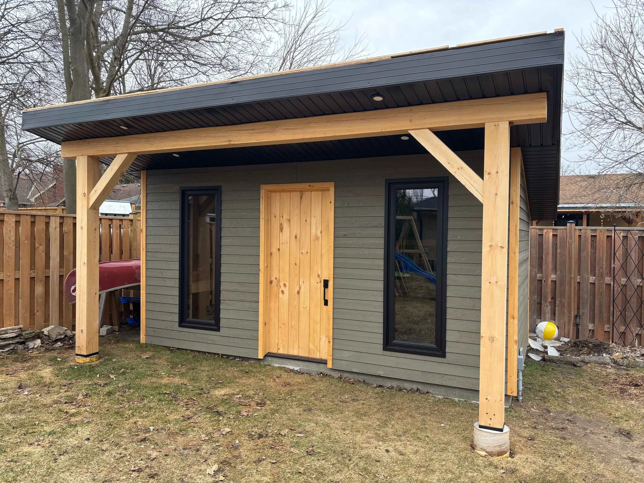 A small detached building or shed with gray siding, black framed windows, and a new wooden door, under construction with a partial roof structure and supporting posts. There is a wooden fence surrounding the yard and some trees in the background.