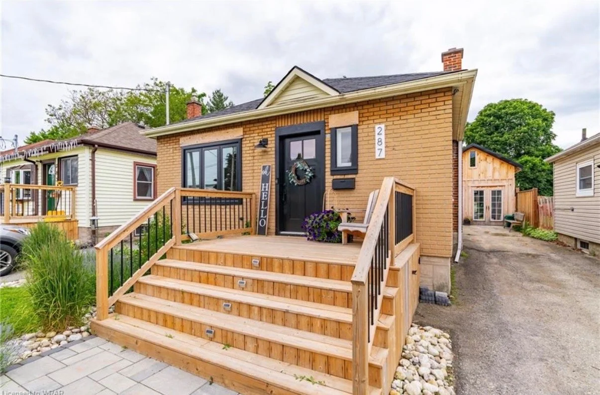 Newly renovated brick house with a wooden front porch, black door with a holiday wreath, and a small sign saying "hello." There are purple flowers on the porch and a bench in the corner. The house has a side yard with a gravel driveway and neighborin