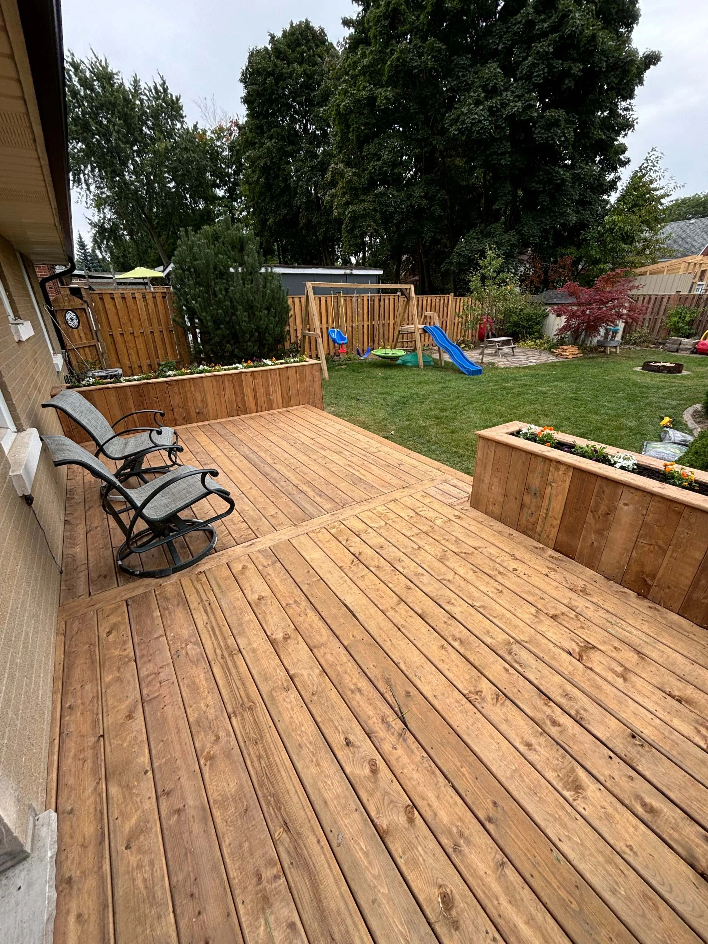 A backyard patio with a wooden deck, outdoor chairs, a grassy area with a playset including a slide, swings, and a small fire pit, enclosed by a wooden fence with trees in the background.