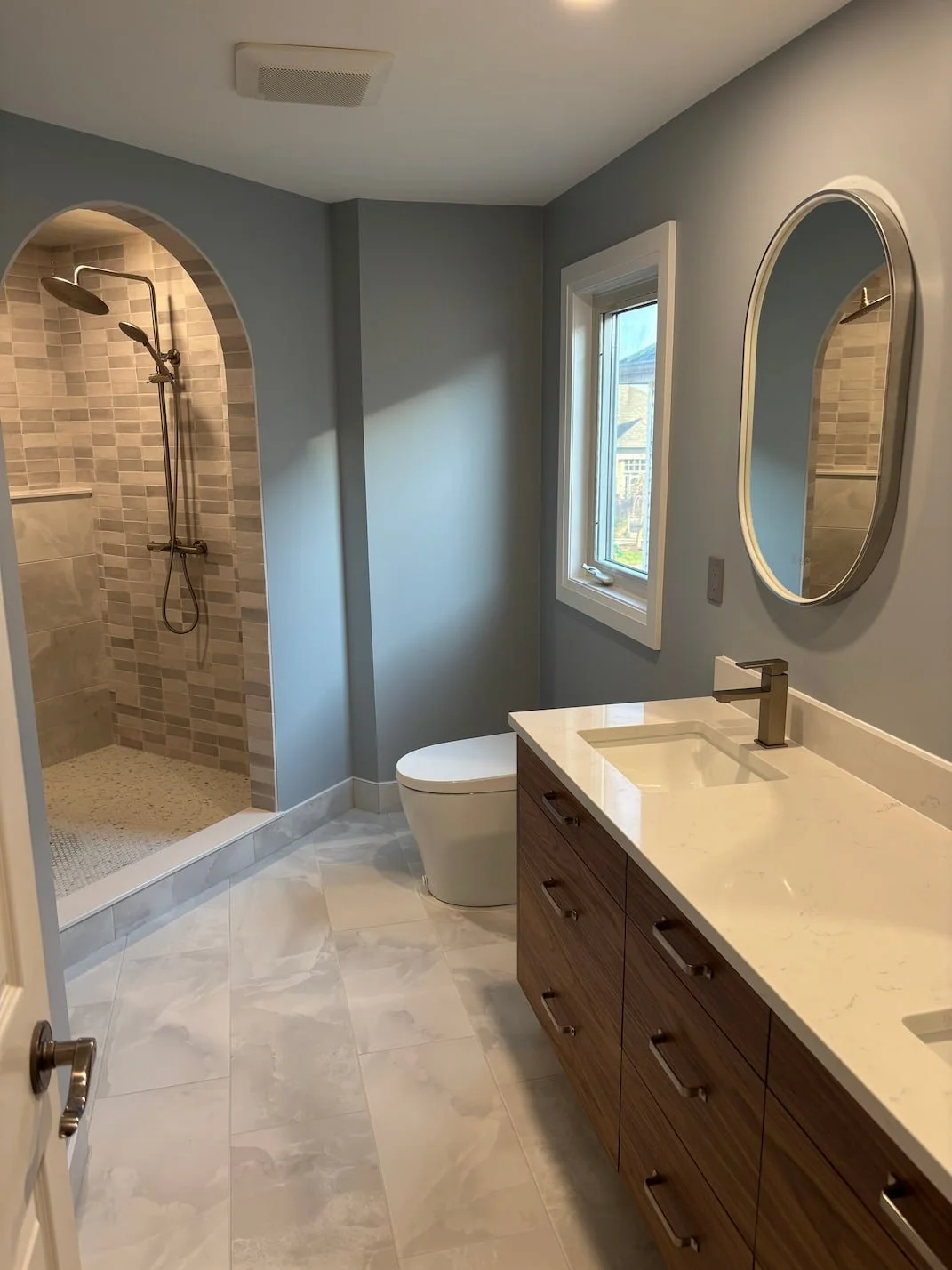 Modern bathroom with walk-in shower, white toilet, marble countertop vanity, oval mirror, and window with a view outside.