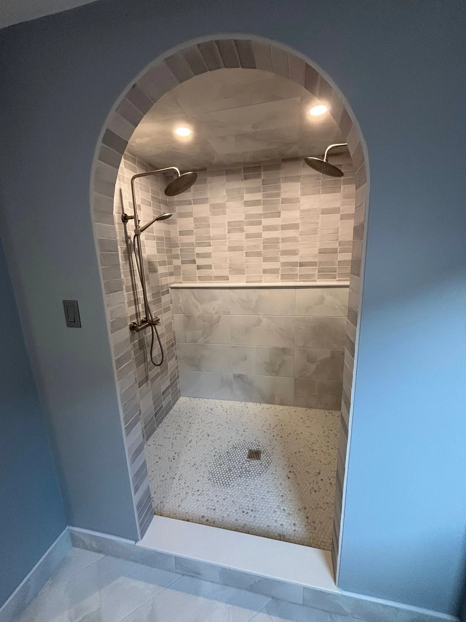 A modern walk-in shower with two rainfall showerheads on the ceiling, tiled walls in beige and light gray, and a textured beige tile floor with a drain in the center.