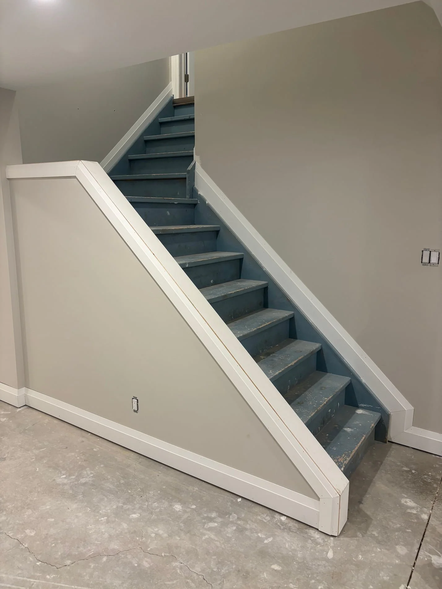 Interior staircase with unfinished blue steps and white framing, next to a beige wall with electrical outlets, on a grey concrete floor.